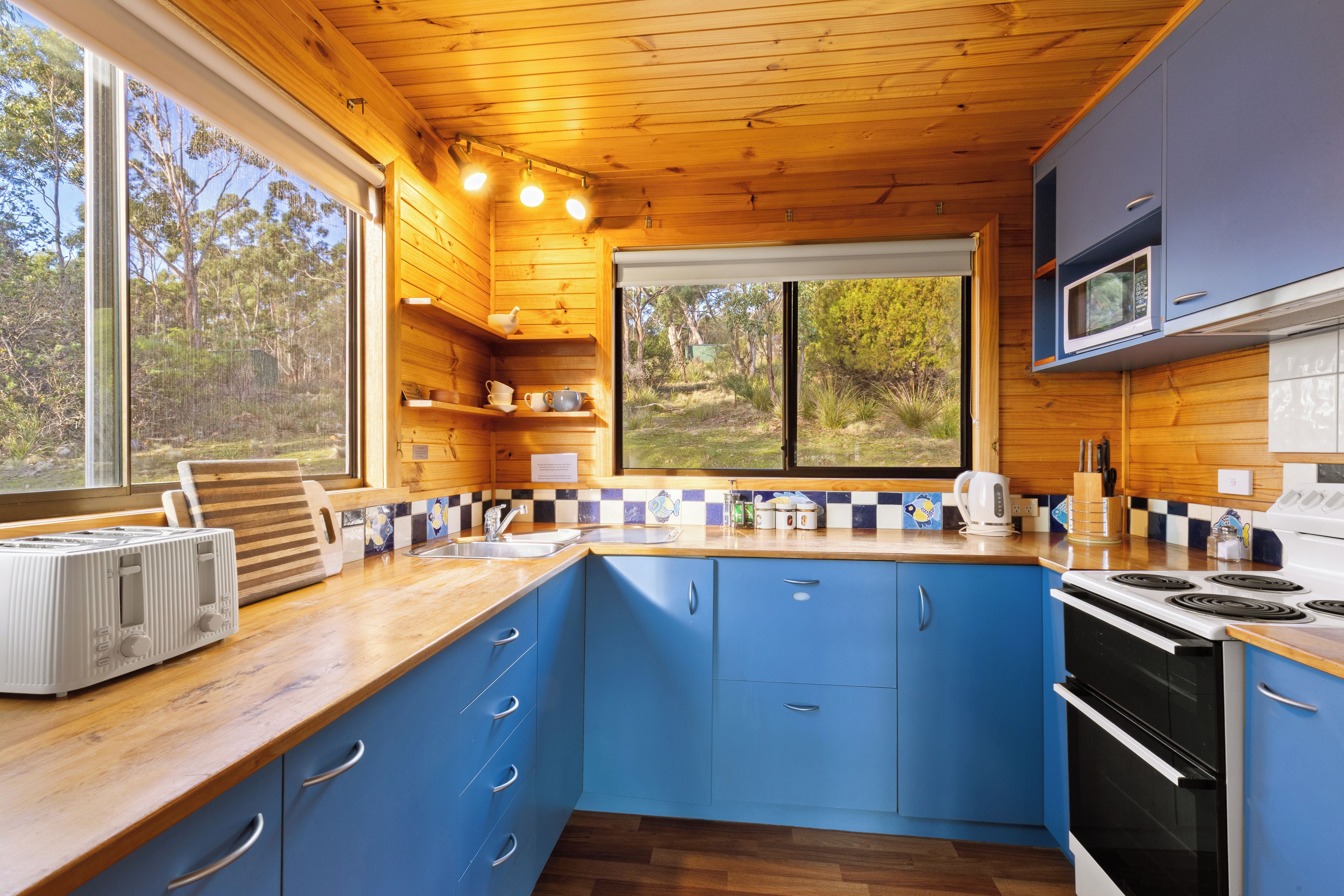 A compact kitchen with wooden cabinets, a microwave, a stove, and a window overlooking a natural landscape.