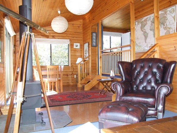 Interior de una cabaña de madera con un sillón de cuero, una mesa de comedor con sillas, una chimenea y un gran mapa mundial en la pared.