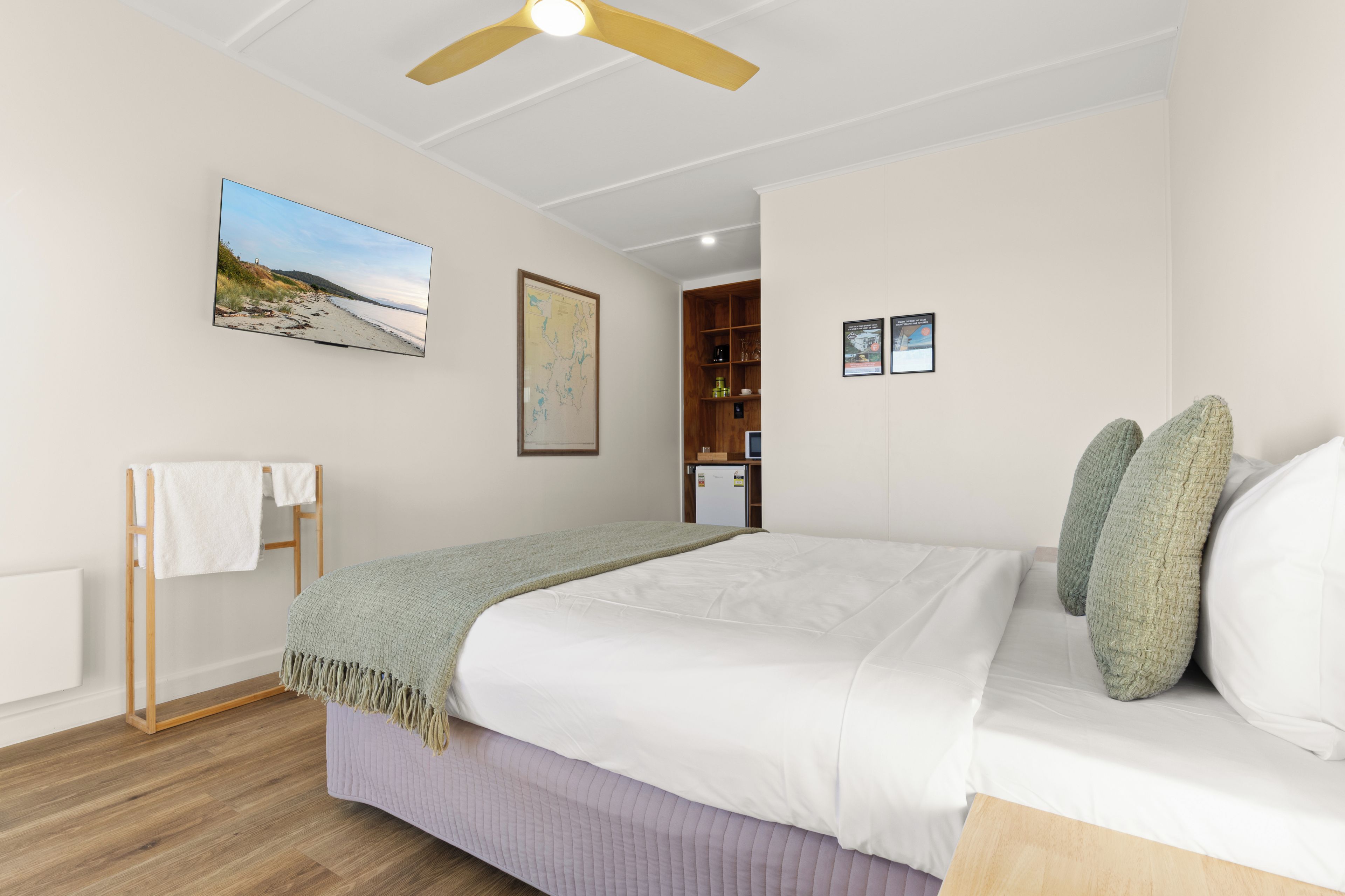 Un dormitorio con una cama, un ventilador de techo, un televisor montado en la pared y un estante de madera con objetos.