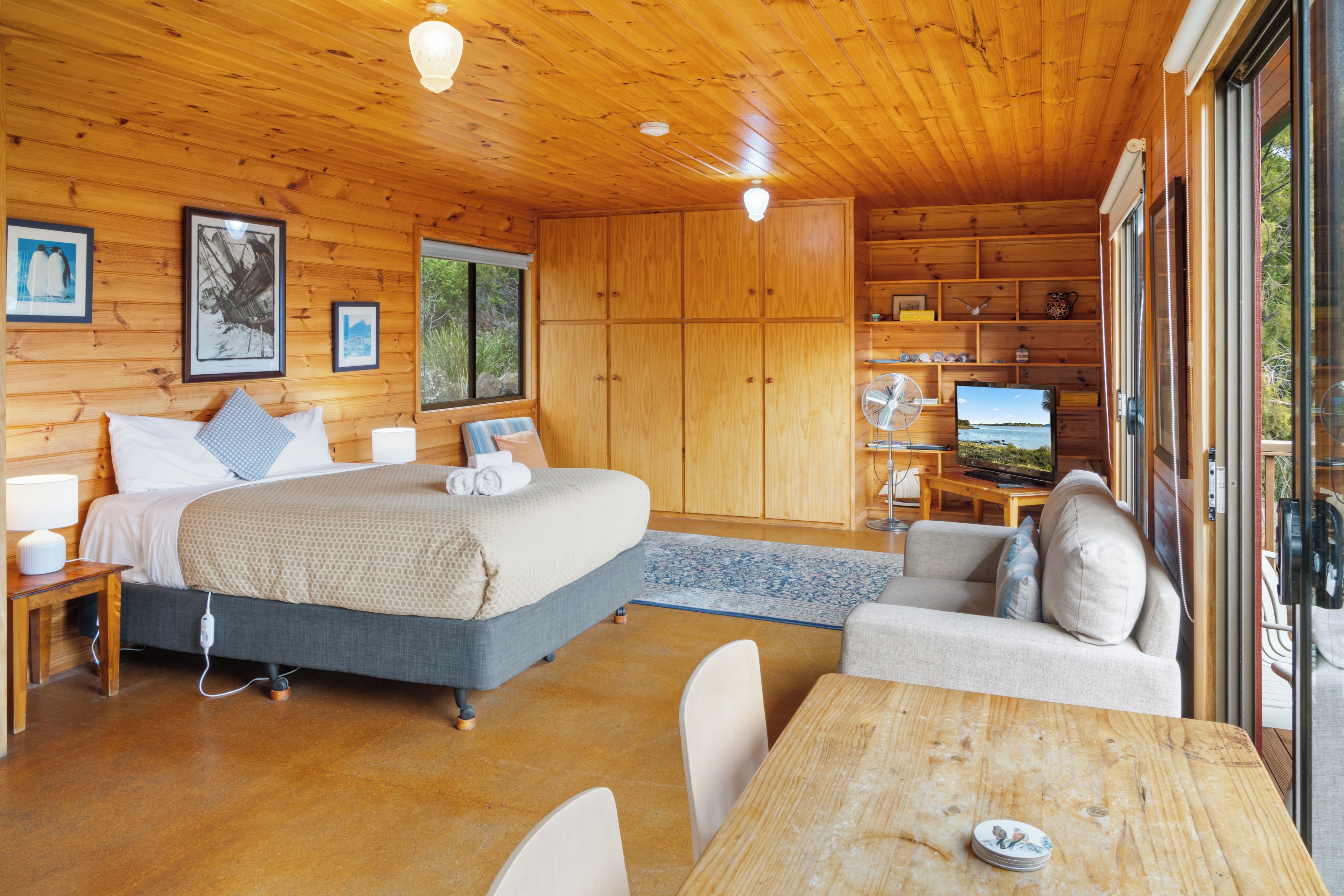 A wooden cabin interior with a bed, bedside tables, a TV, and a dining area.