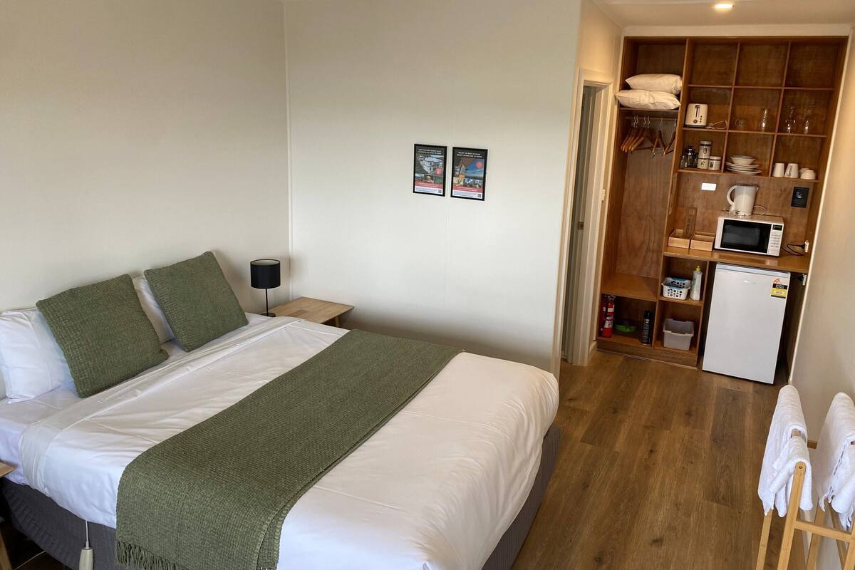 Une chambre comprenant un lit, un ventilateur de plafond, une table de chevet, une lampe fixée au mur et une kitchenette équipée d’un four à micro-ondes et d’un réfrigérateur.