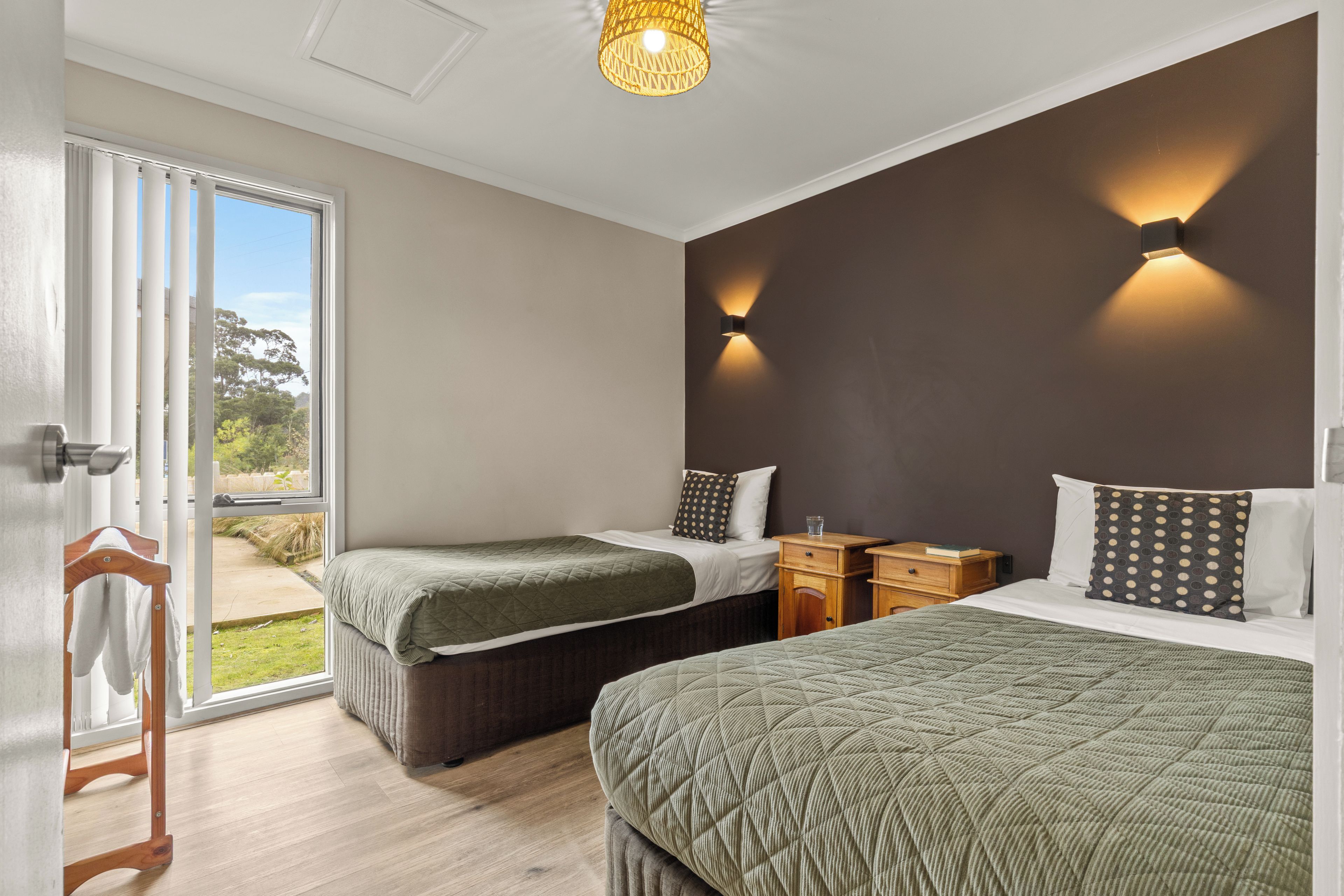 A bedroom with two beds, a wooden nightstand, a wall-mounted light, and a view through a sliding door.