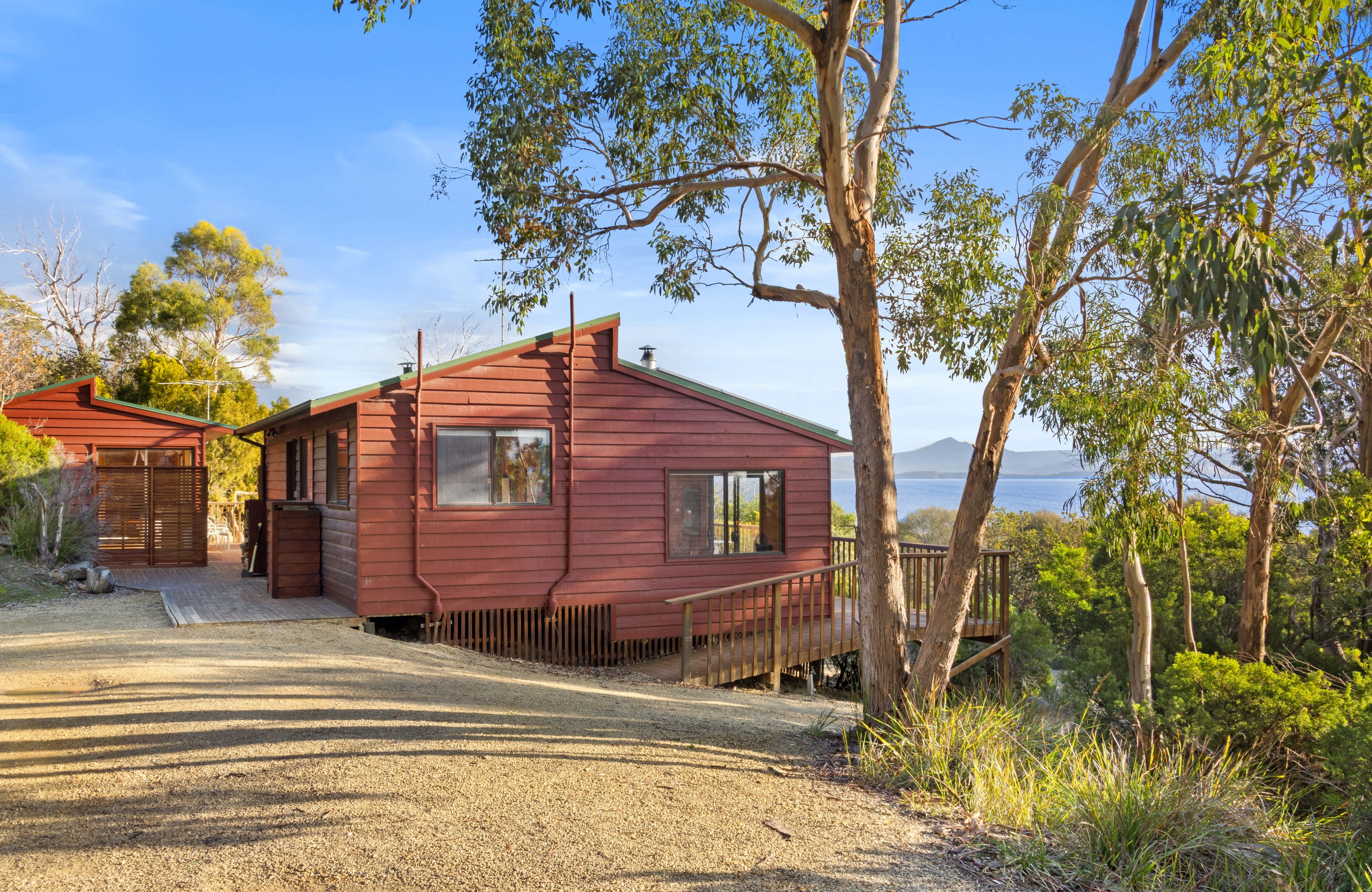 A red cabin with a deck, surrounded by trees and a gravel driveway.