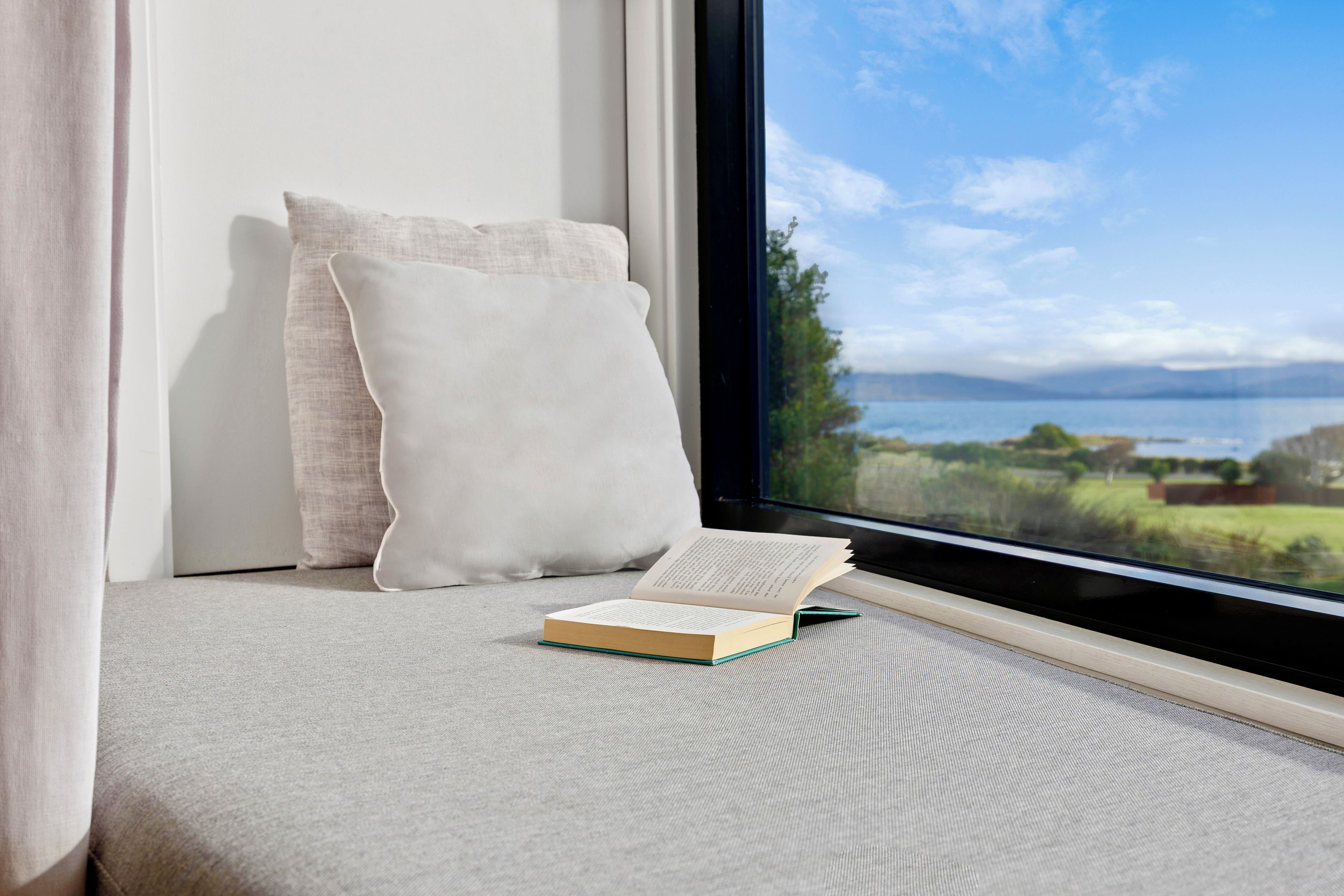 A window seat with a cushion, a book, and a view of a lake and mountains.