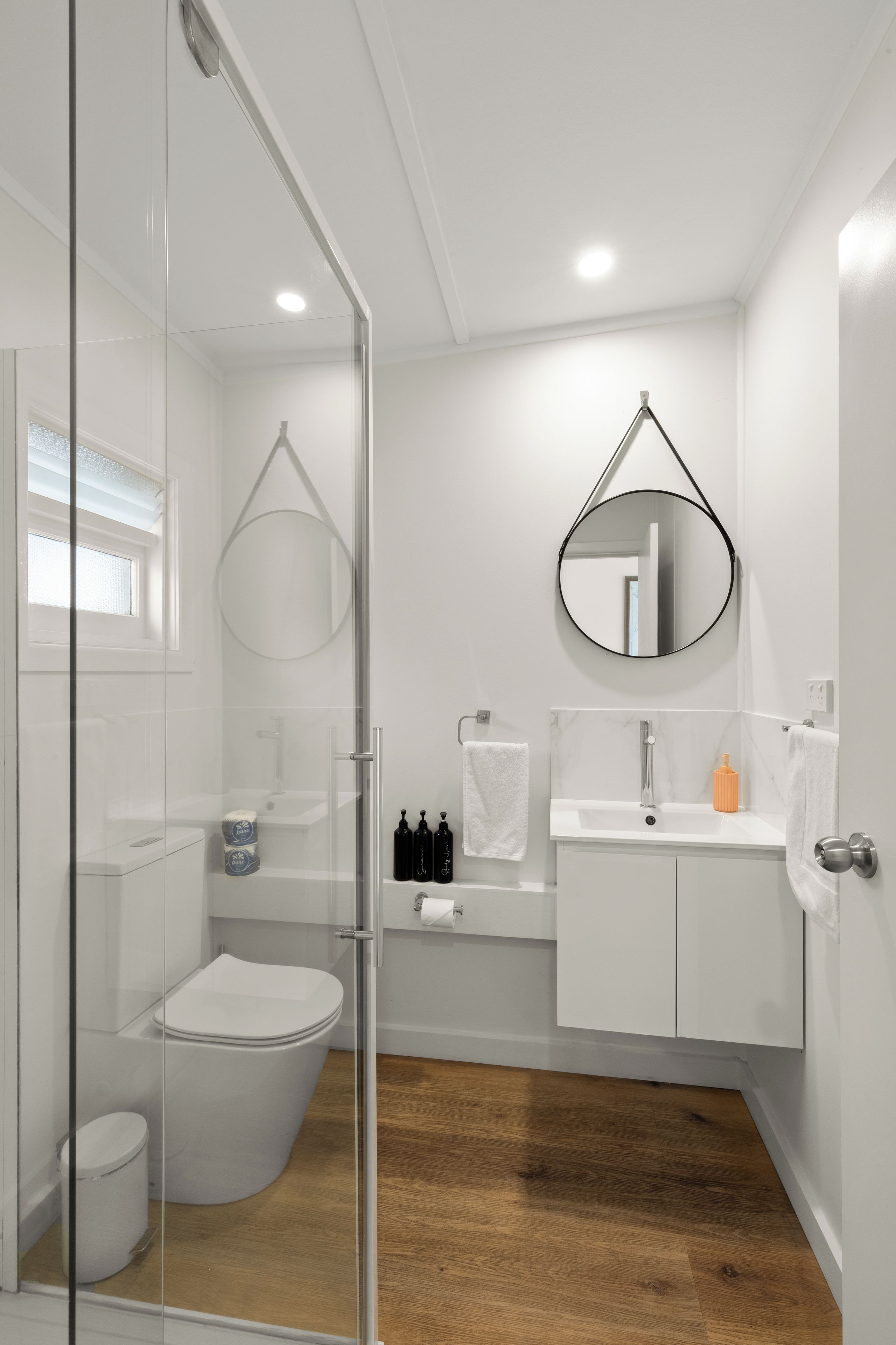 Un baño moderno con cabina de ducha de vidrio, inodoro, lavamanos con espejo y suelo de madera.