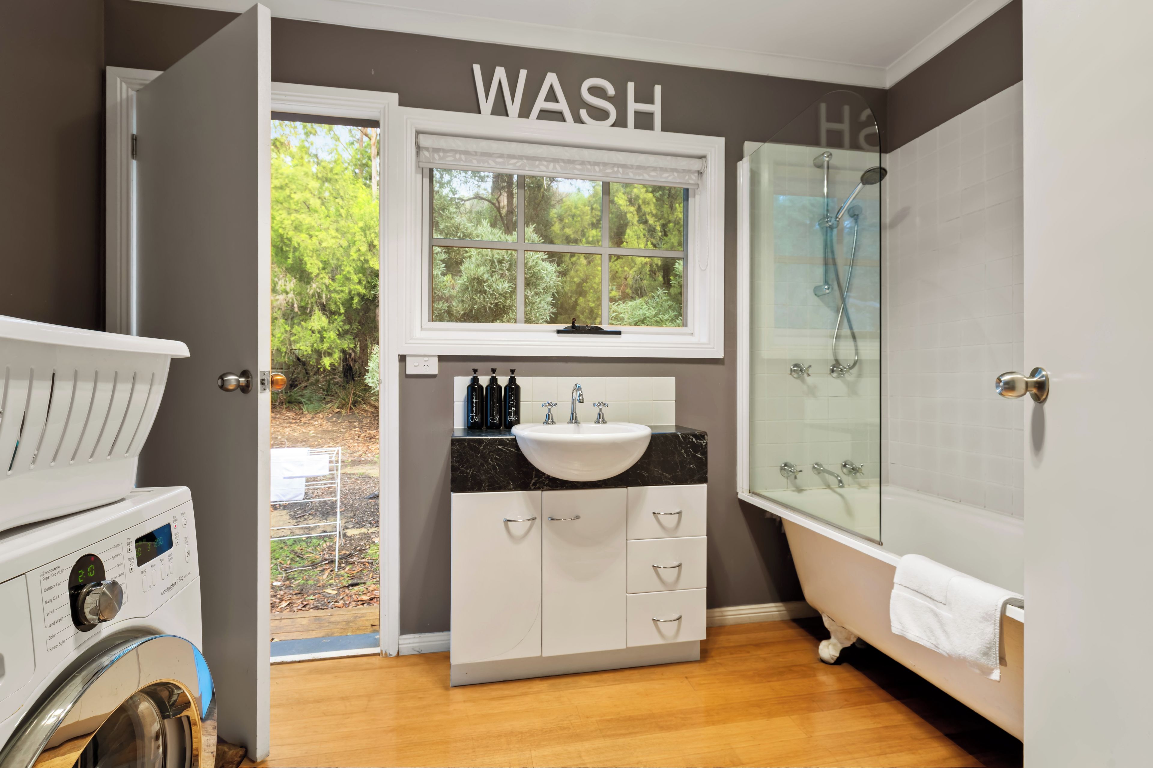 A modern bathroom with a built-in washer and dryer, a large window with a view of greenery, a vanity with a sink and mirror, and a walk-in shower.