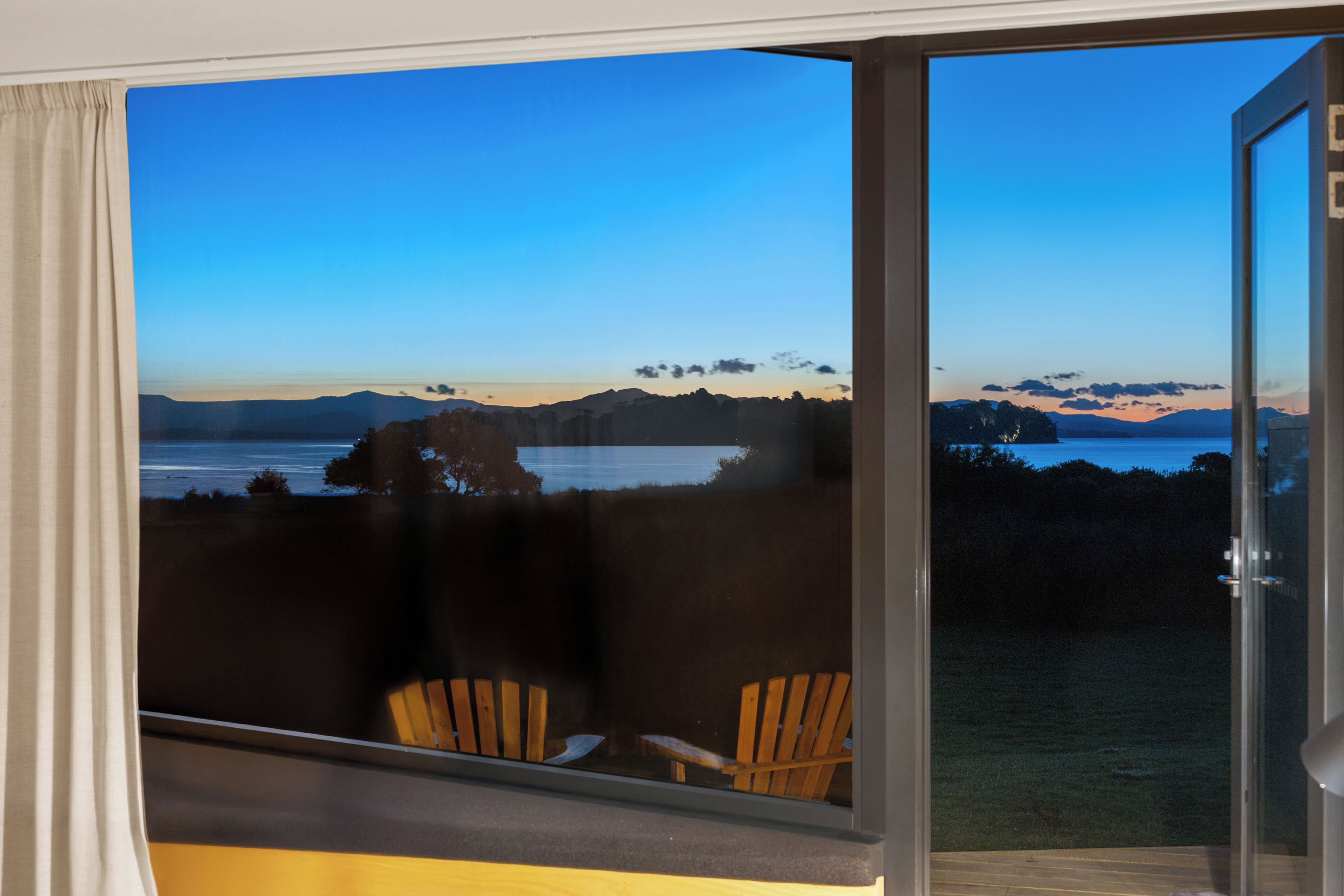 A room with a view of a lake and mountains, two wooden chairs, and a glass door.