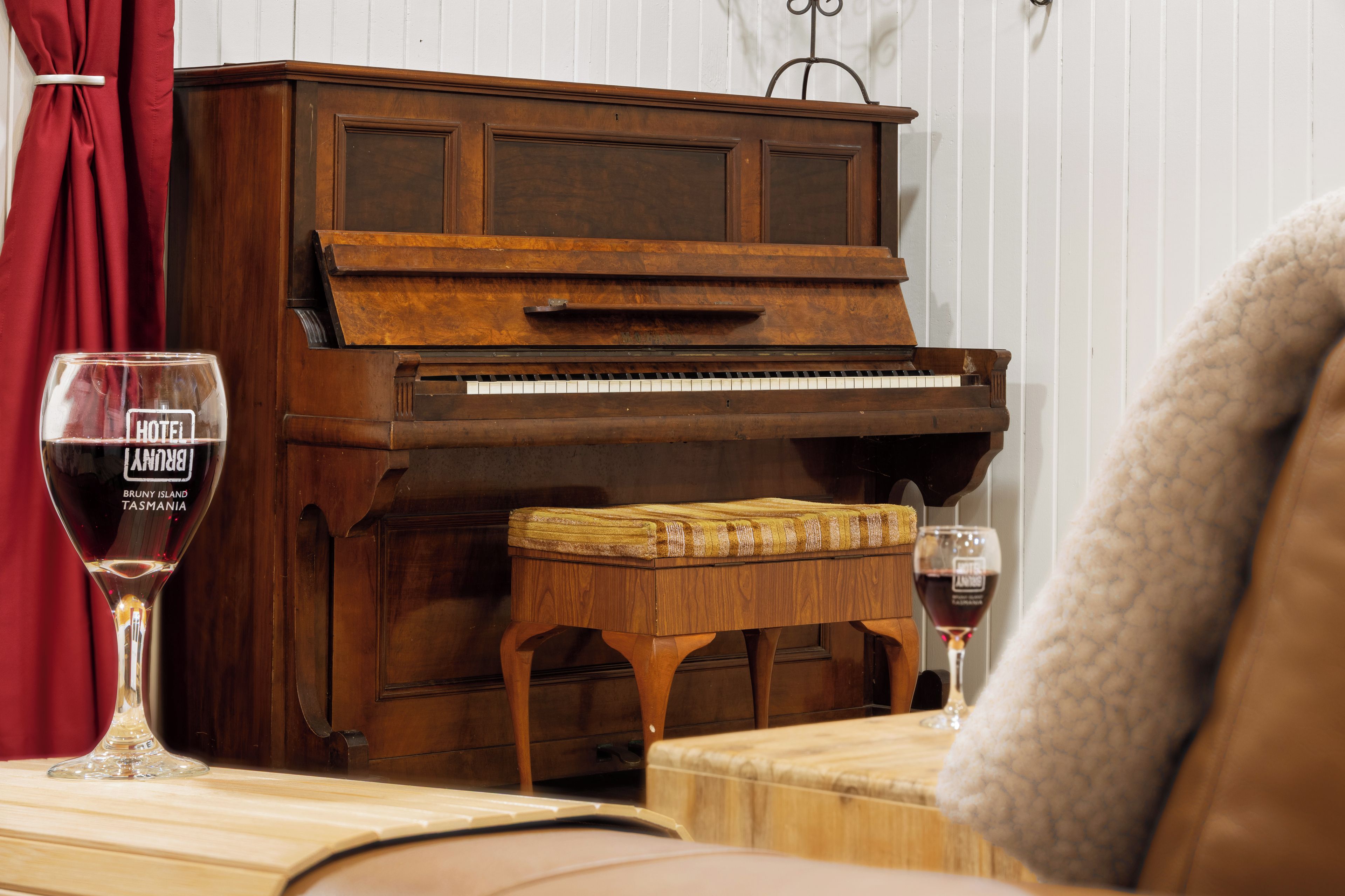 A wooden piano with a bench in front of it, a glass of red wine on a table, and a red curtain in the background.