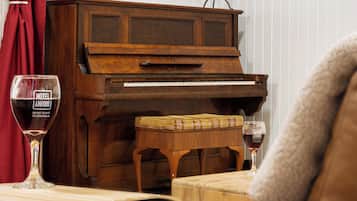 A wooden piano with a bench in front of it, a glass of red wine on a table, and a red curtain in the background.