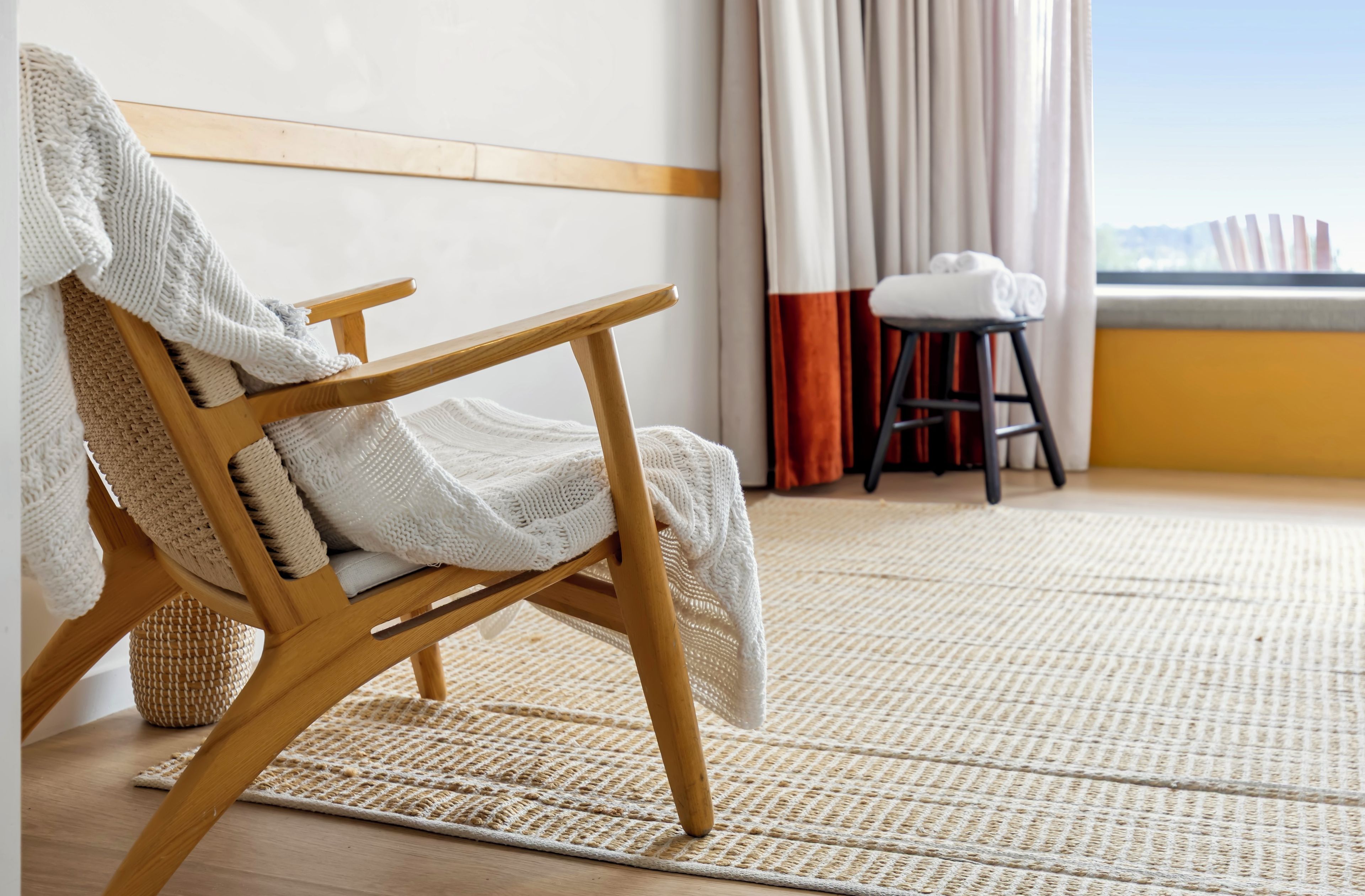 A wooden chair with a white knitted throw, a woven rug, and a view of the outdoors through a window with curtains.