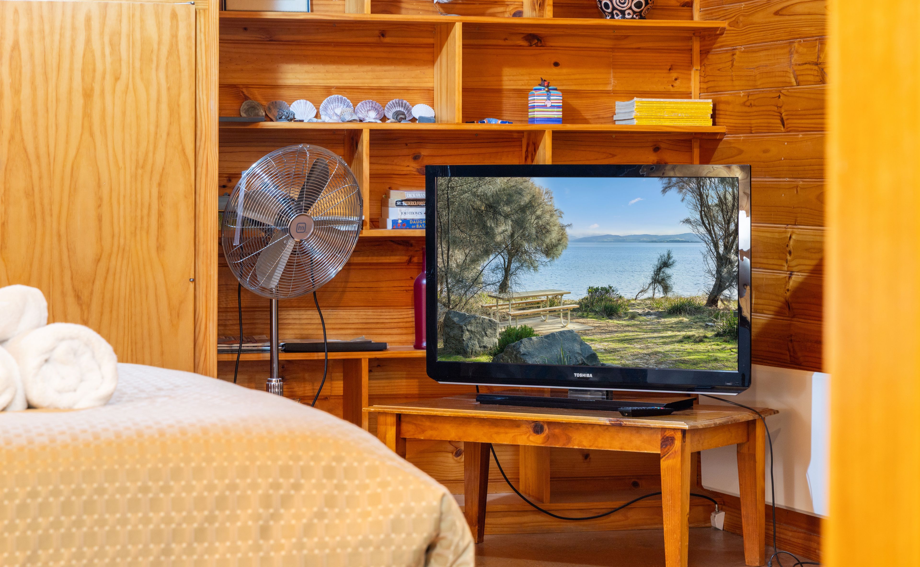 Una habitación con interior de madera, una cama con edredón blanco, un televisor sobre un mueble de madera mostrando una vista panorámica y un ventilador en el suelo.