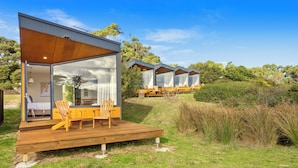 A modern outdoor deck with wooden furniture, a glass door leading to a room, and a view of a row of similar structures in the background. - Bruny Island Escapes and Hotel Bruny (Alonnah)