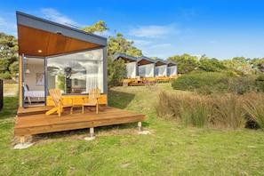 A modern outdoor deck with wooden furniture, a glass door leading to a room, and a view of a row of similar structures in the background. - Bruny Island Escapes and Hotel Bruny (Alonnah)