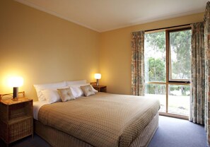 A bedroom with a large bed, two bedside lamps, a window with curtains, and a view of trees. - Bruny Island Escapes and Hotel Bruny (Alonnah)