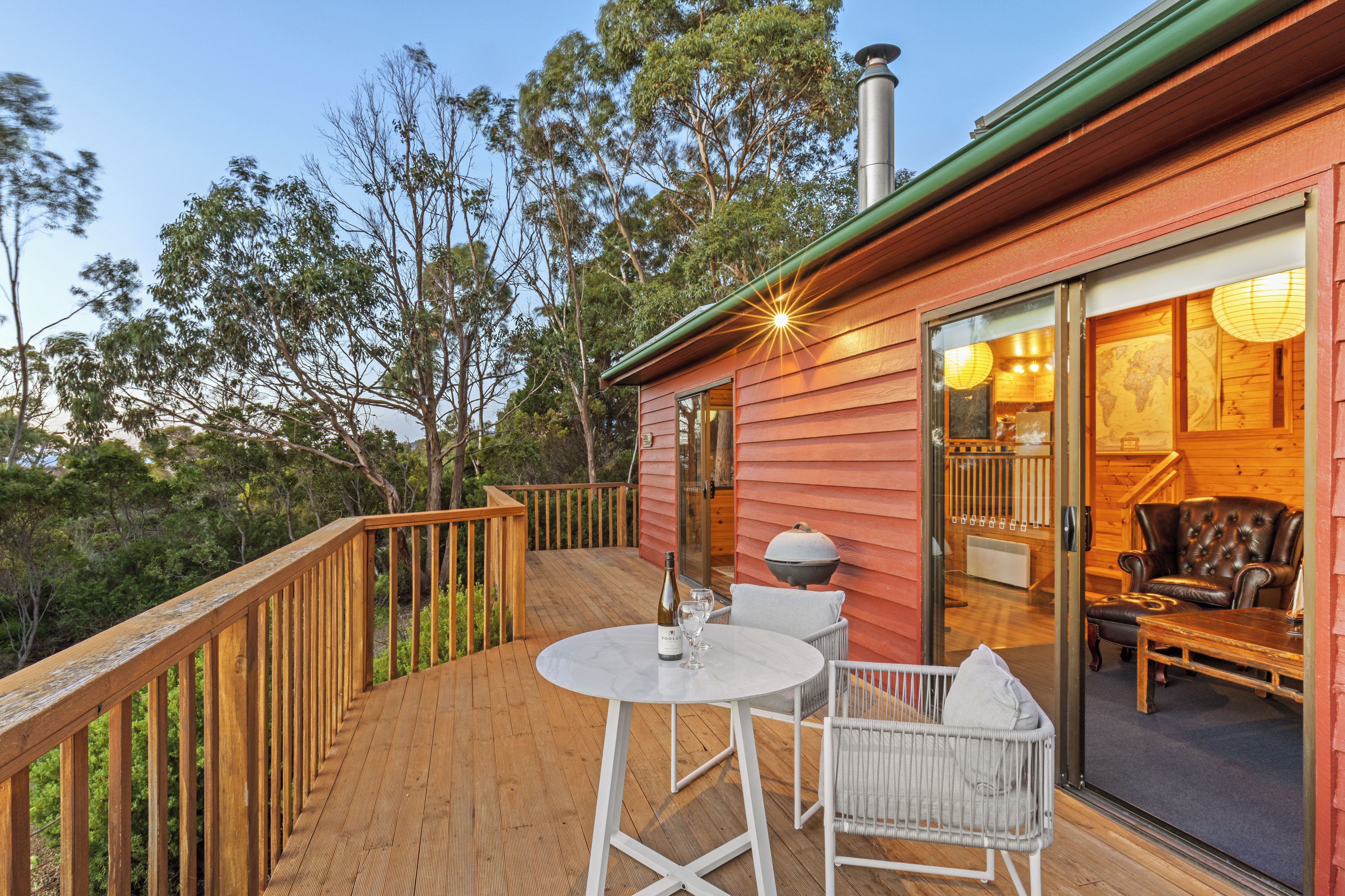 Une terrasse en bois avec une table et des chaises, une porte vitrée donnant sur un espace intérieur équipé d’un fauteuil et d’une lampe.