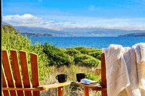 A wooden chair with a black mug and a newspaper, a white knitted blanket, and a view of a body of water with mountains in the distance. - Bruny Island Escapes and Hotel Bruny (Alonnah)