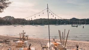 A beachside dining setup with tables and chairs, overlooking a calm sea with boats and hills in the background. - Samui Honey Cottages Beach Resort (Koh Samui)