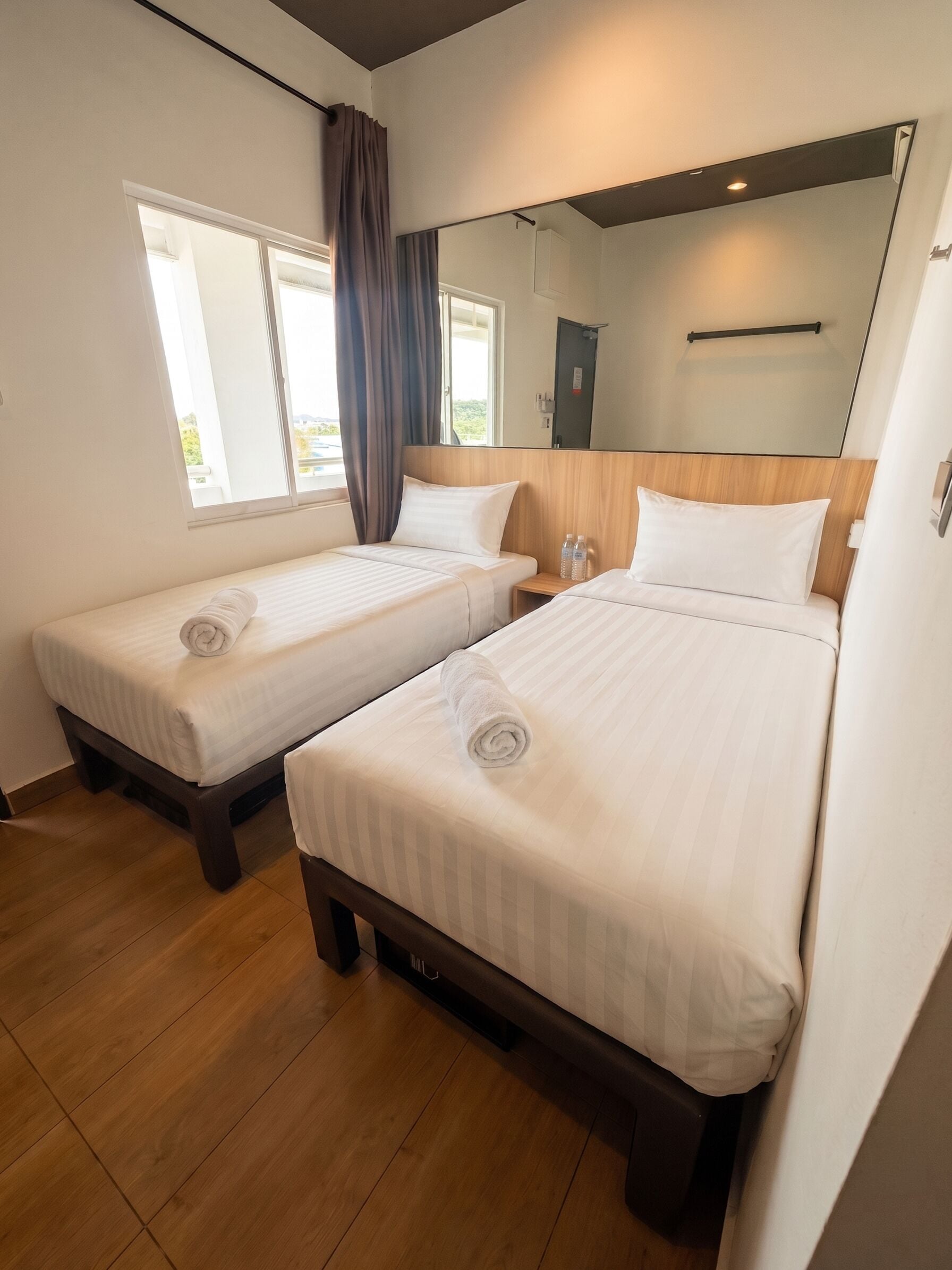 Habitación de hotel con dos camas individuales, suelo de madera, un ventanal con cortinas y un espejo en la pared.