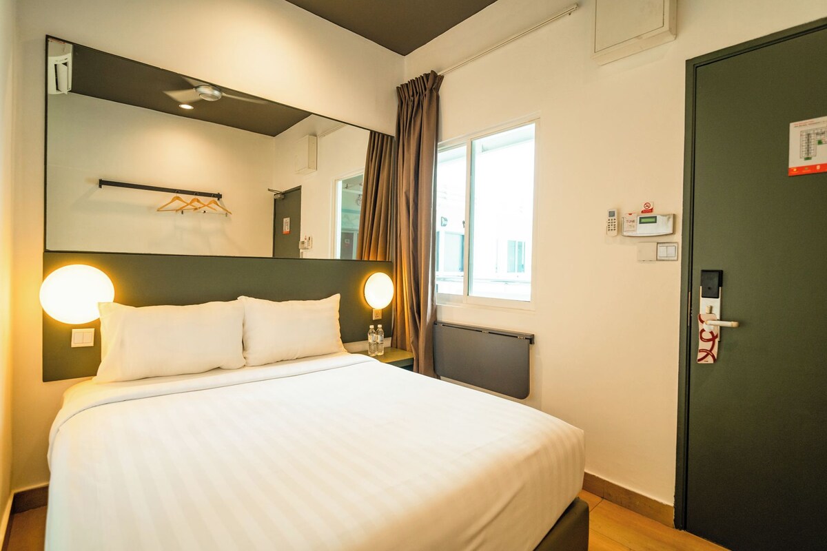 A hotel room with a bed, bedside lamps, a window with curtains, a door with a handle, and a thermostat on the wall.