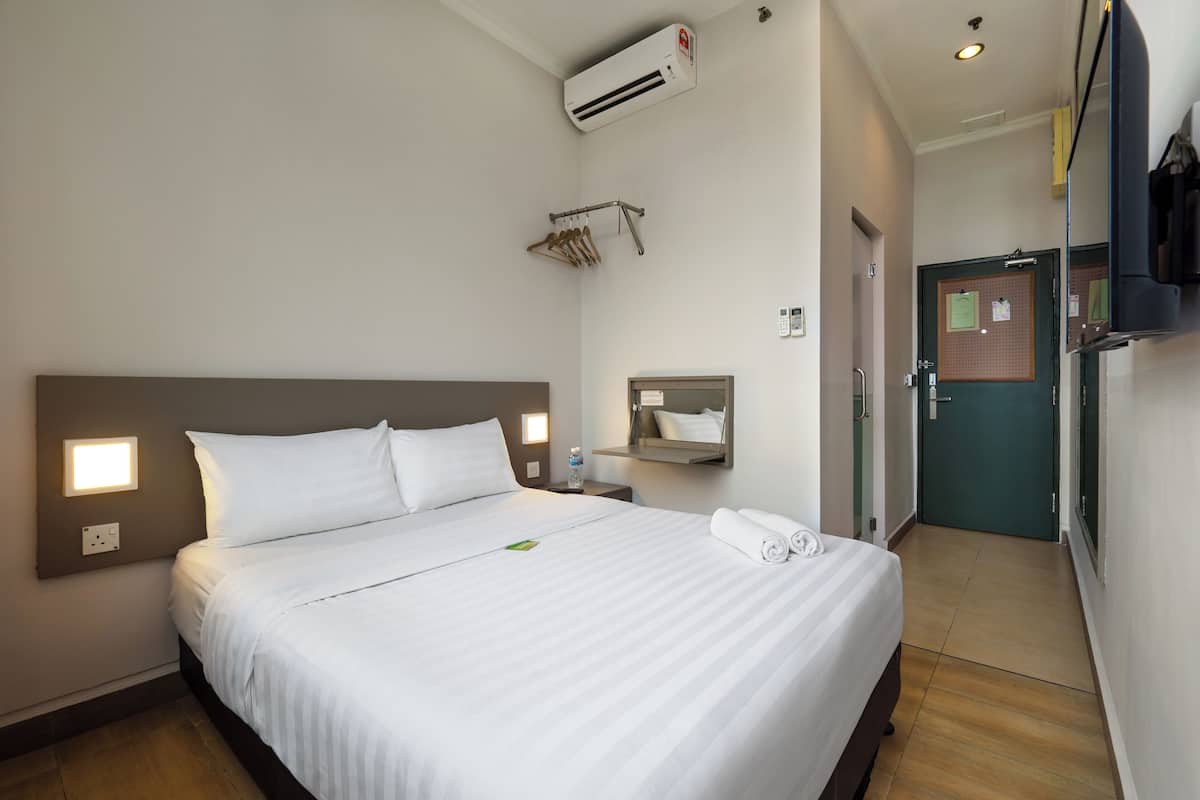 A hotel room with a large bed, bedside table, wall-mounted air conditioning, a mirror, a door with a bulletin board, and a small window.
