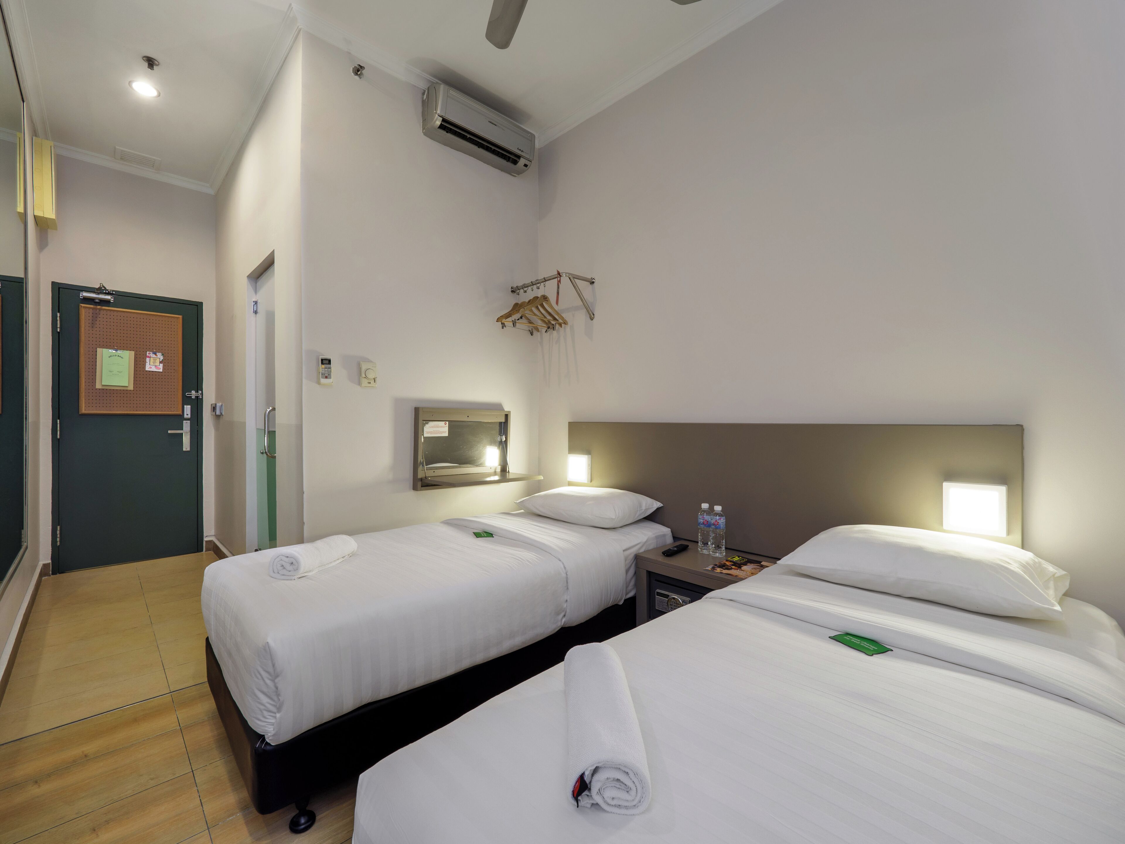 Une chambre d’hôtel avec deux lits, une table de chevet avec une bouteille d’eau, un climatiseur fixé au mur, une porte avec un tableau d’affichage et une petite fenêtre.
