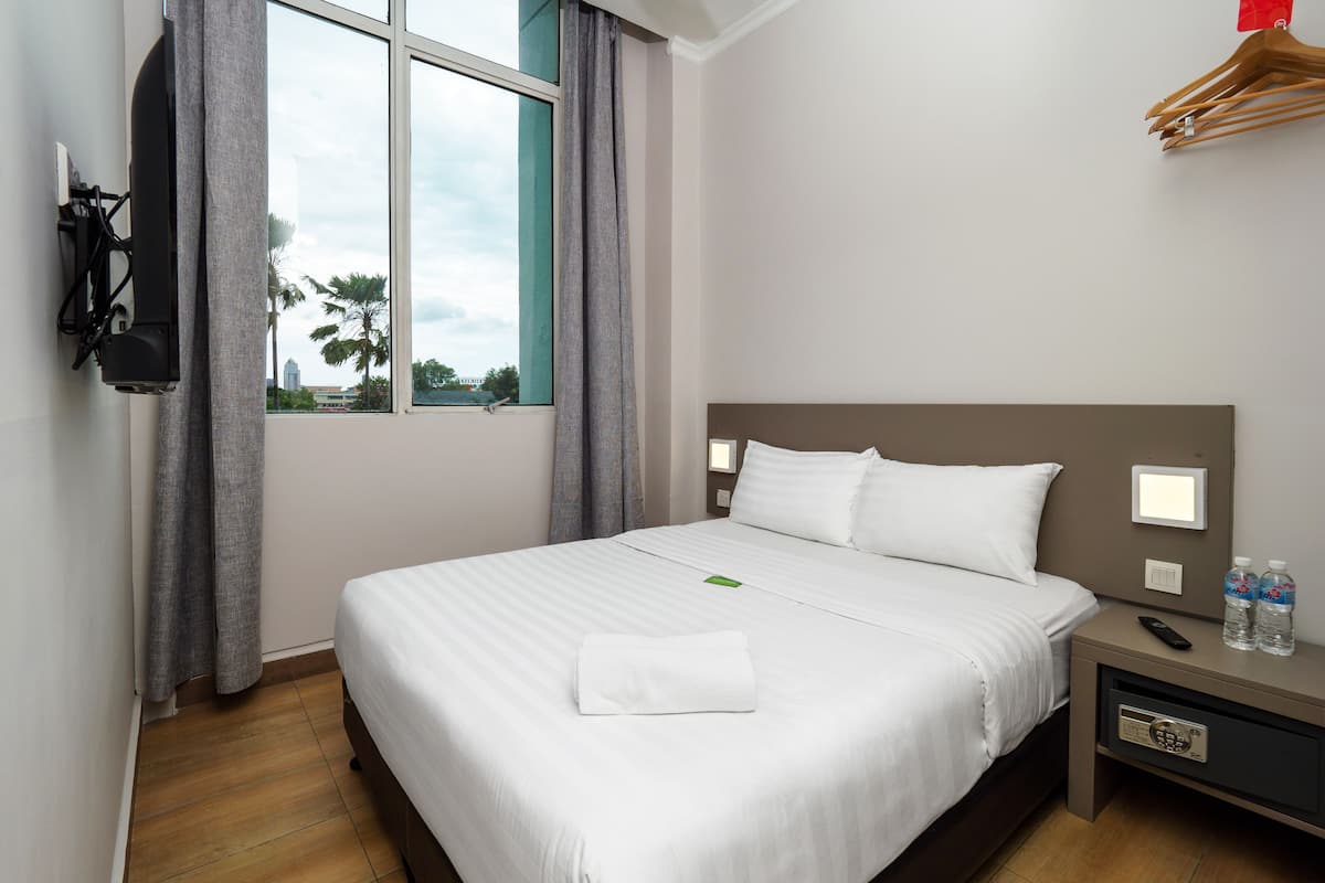A hotel room with a large bed, a TV mounted on the wall, a bedside table with a remote control, and a window with curtains.
