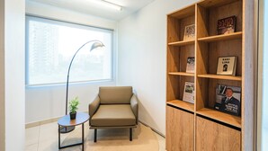 A modern office space with a beige chair, a small round table with a potted plant, and a wooden bookshelf with various items including books and framed pictures. - NIO Hotel (George Town)