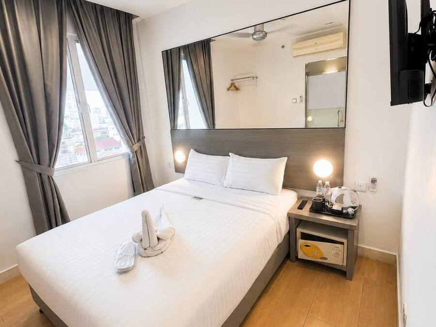 A hotel room with a bed, a bedside table with a lamp, a flat-screen TV mounted on the wall, and a large mirror above the bed.