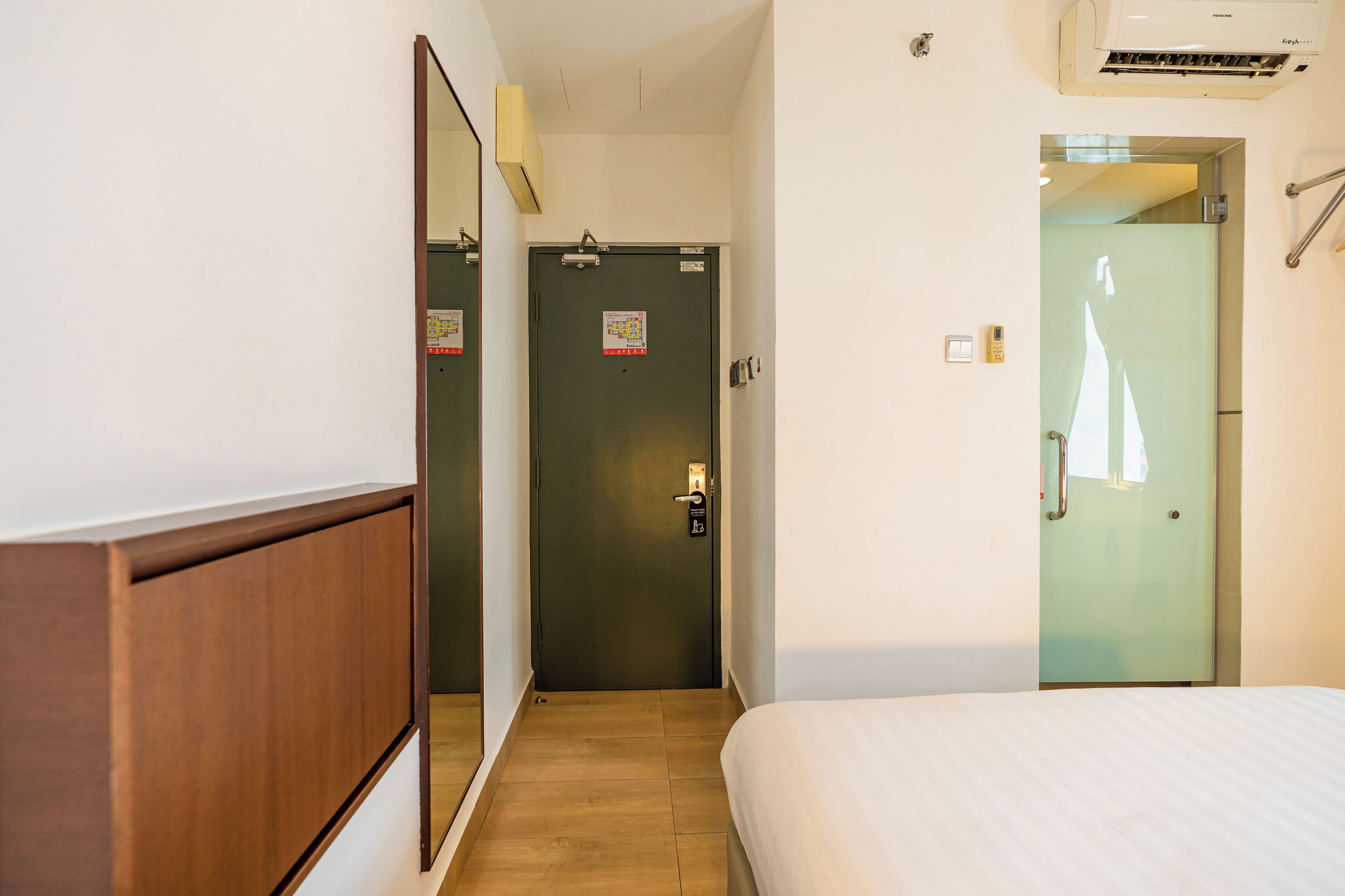 A hotel room with a wooden wardrobe, a bed with white bedding, a door with a mirror, and a wall-mounted air conditioner.