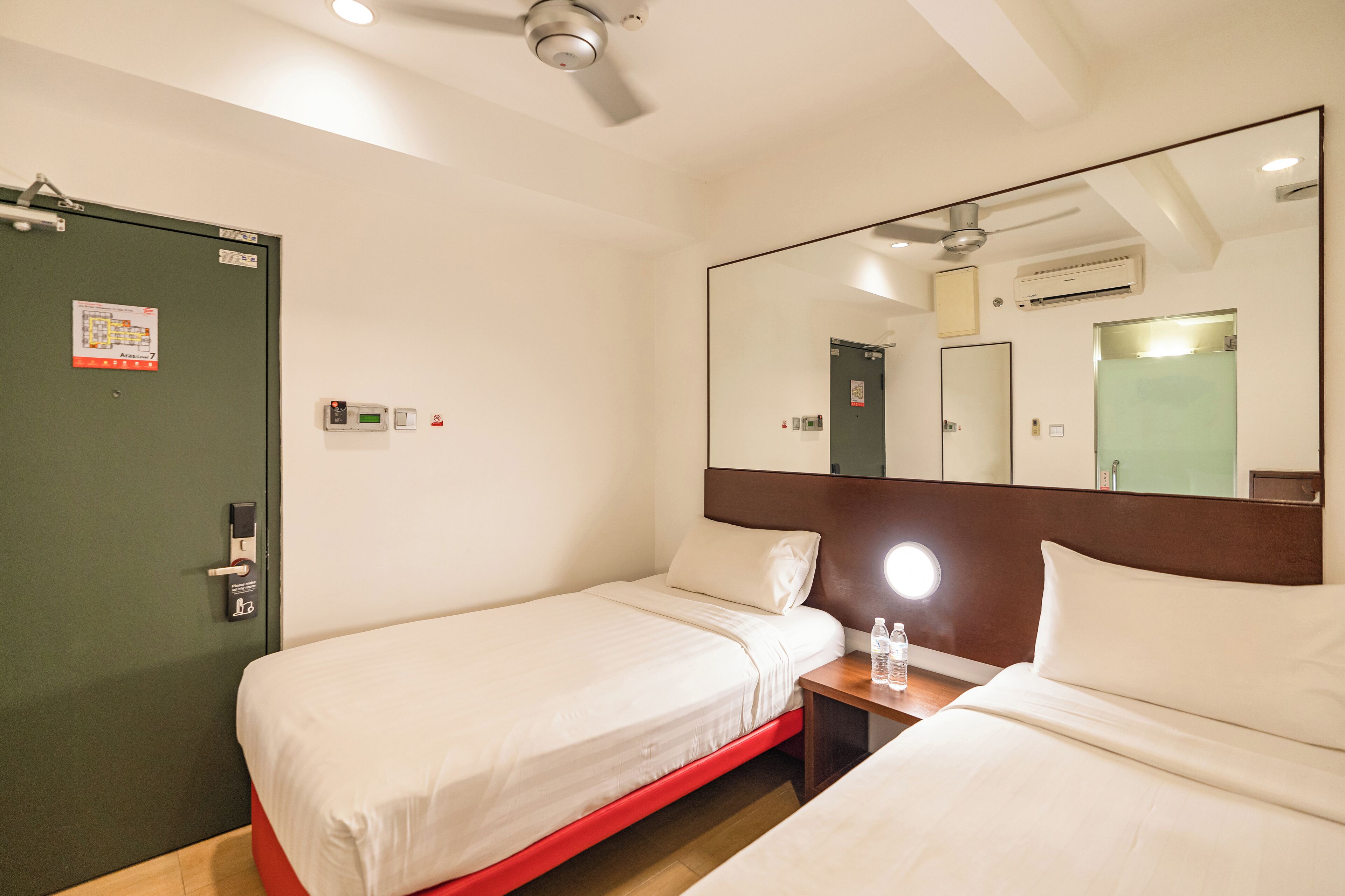 A hotel room with two single beds, a bedside table with a lamp, a large mirror, and a ceiling fan.