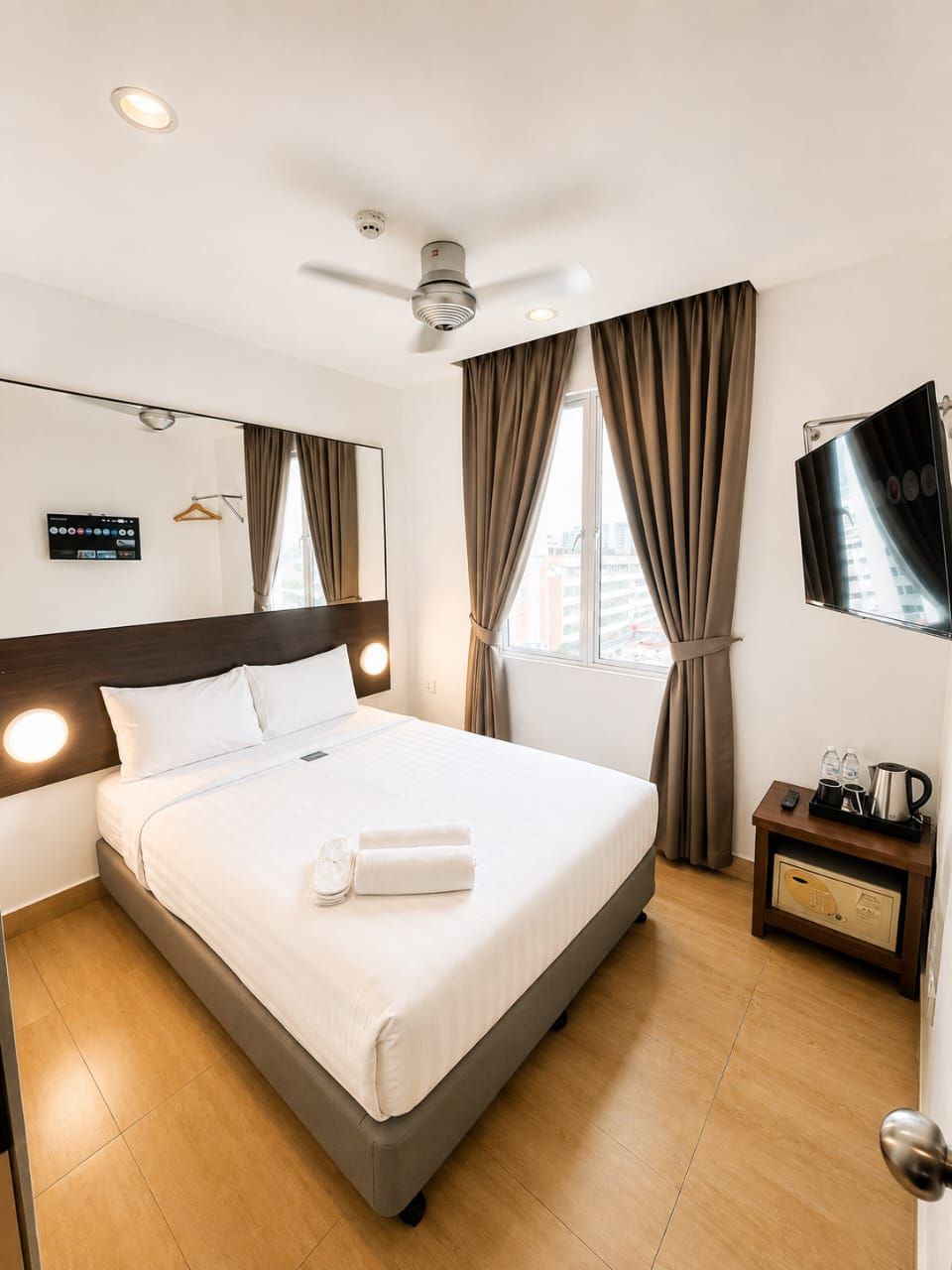 A hotel room with a bed, a bedside table with a remote control, a TV, a ceiling fan, and a window with curtains.