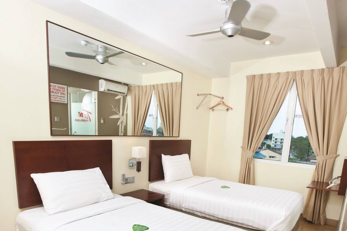 Ein Hotelzimmer mit zwei Betten, einem Deckenventilator, einem Spiegel, einem Fenster mit Vorhängen und Blick nach draußen.