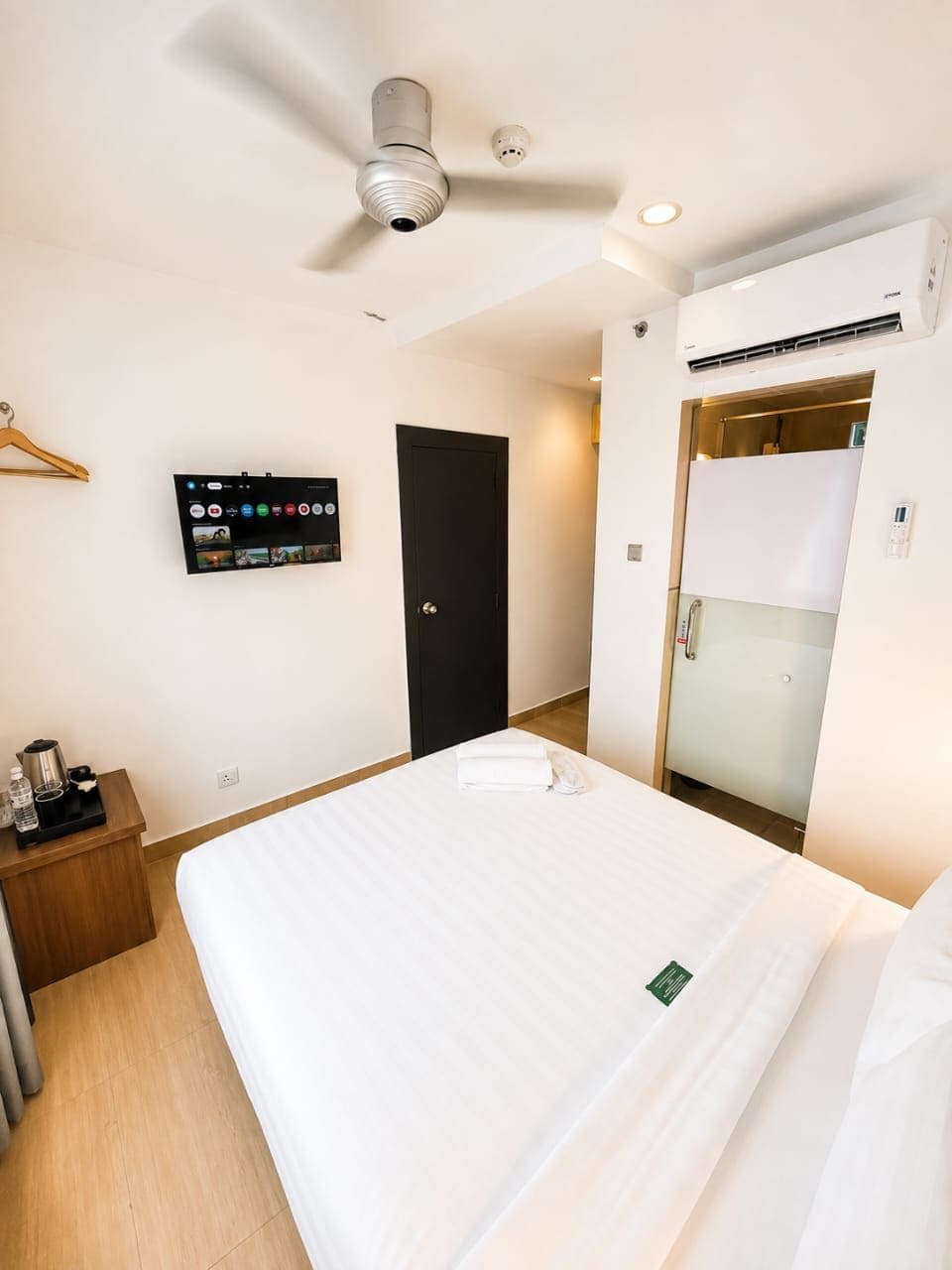 A hotel room with a bed, a television mounted on the wall, a ceiling fan, an air conditioning unit, and a door leading to a bathroom.