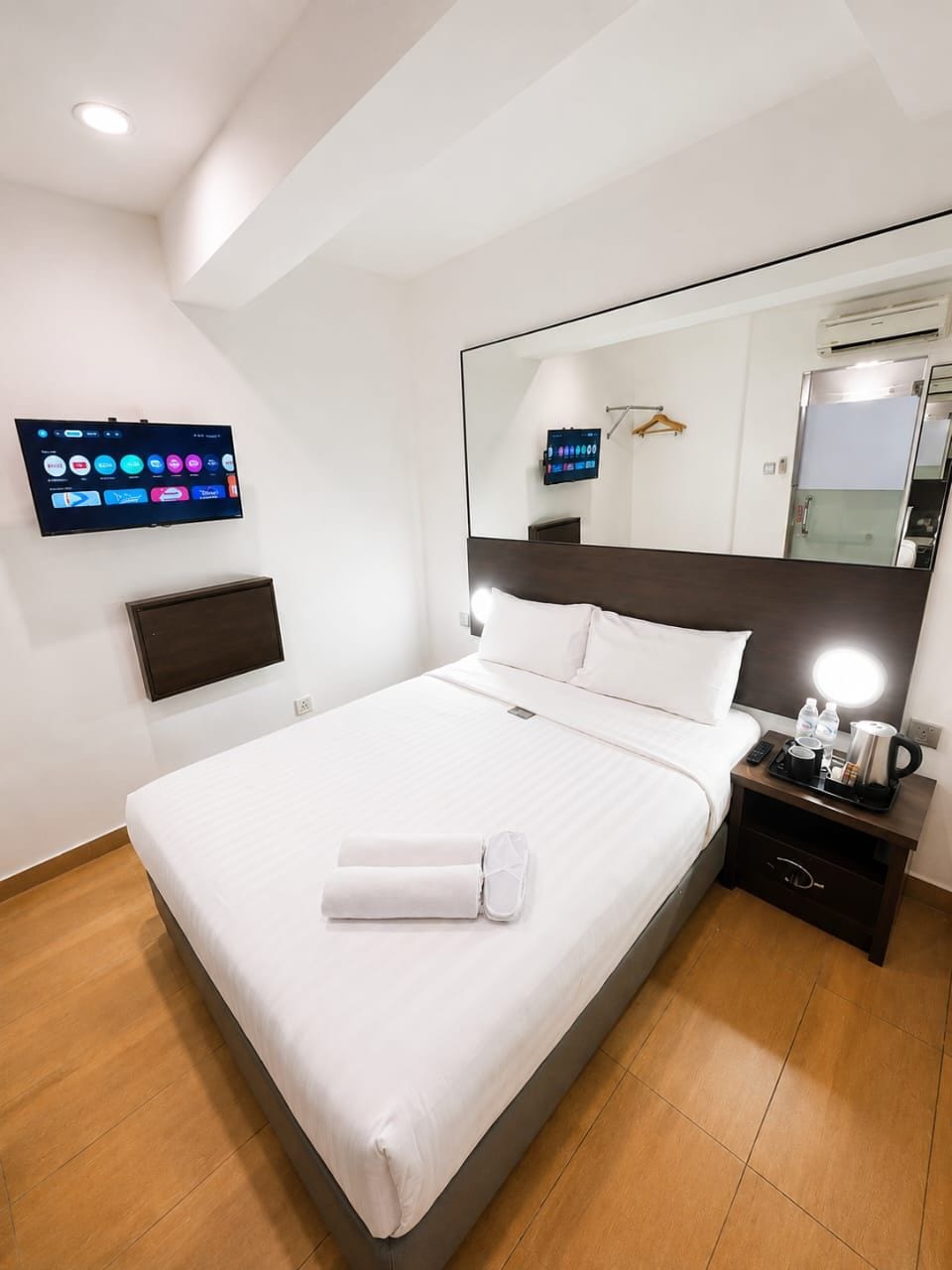 A hotel room with a bed, a flat-screen TV mounted on the wall, a bedside table with a lamp, and a mirror above the bed.