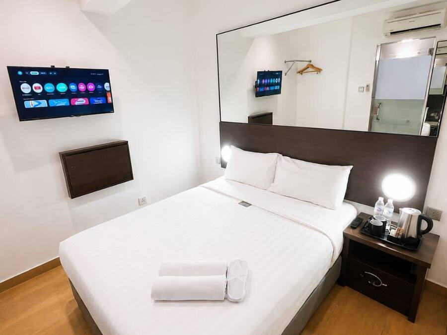 A hotel room with a bed, a flat-screen TV mounted on the wall, a bedside table with a lamp, and a mirror above the bed.