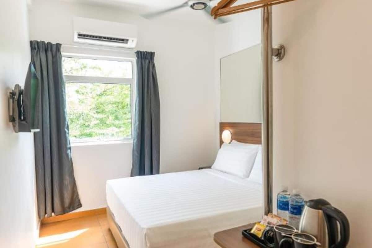 Une chambre d’hôtel de taille réduite, comprenant un lit, un ventilateur de plafond, une fenêtre avec des rideaux, une table de chevet avec une bouilloire et des tasses, ainsi qu’un appareil de climatisation.
