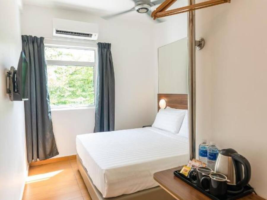 Een compacte hotelkamer met een bed, een plafondventilator, een raam met gordijnen, een nachtkastje met een waterkoker en kopjes, en een airconditioning.
