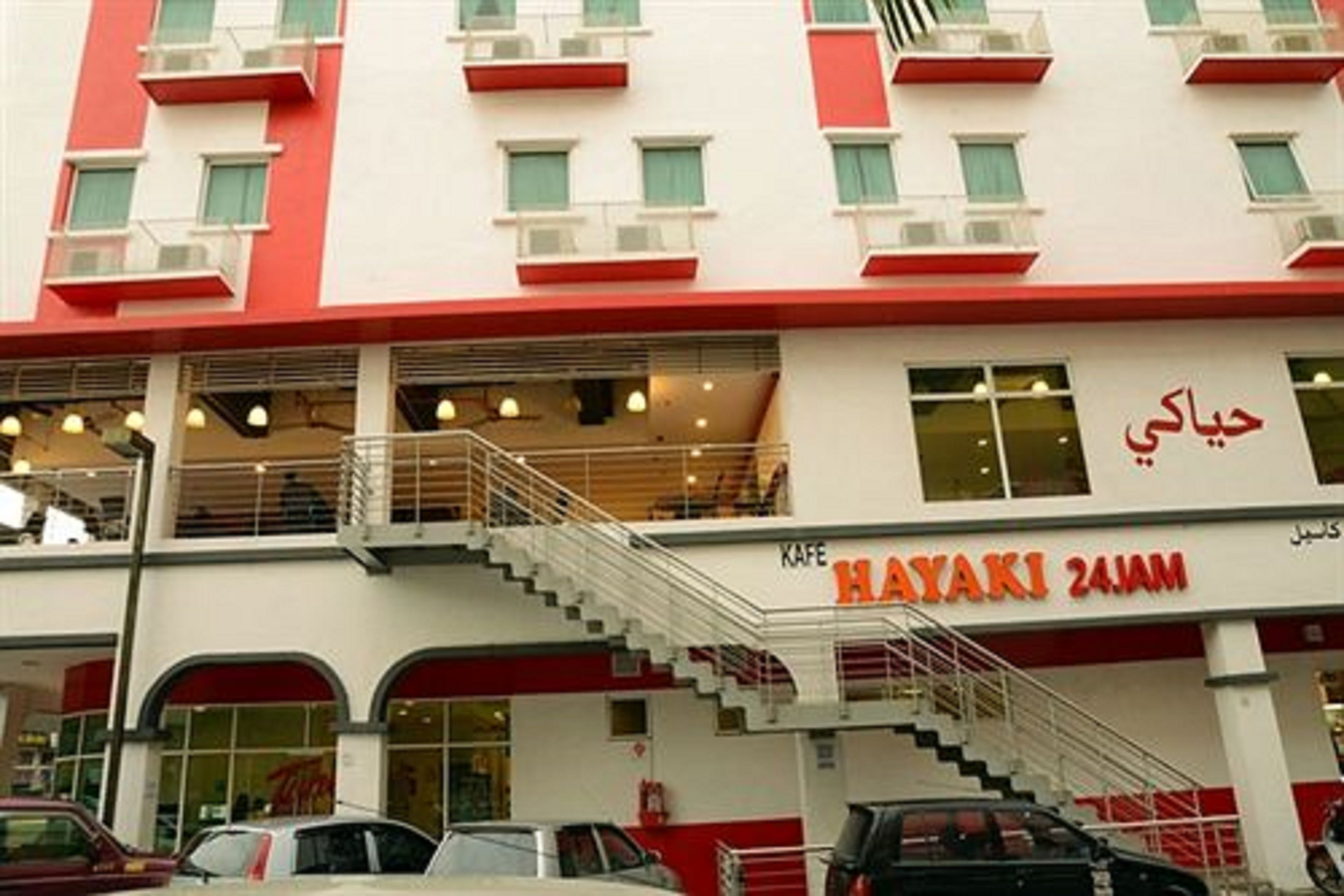 Um edifício com vários andares, fachada vermelha e branca, e uma escadaria que leva ao andar superior, com uma placa indicando o nome "HAYAKI 24AM".