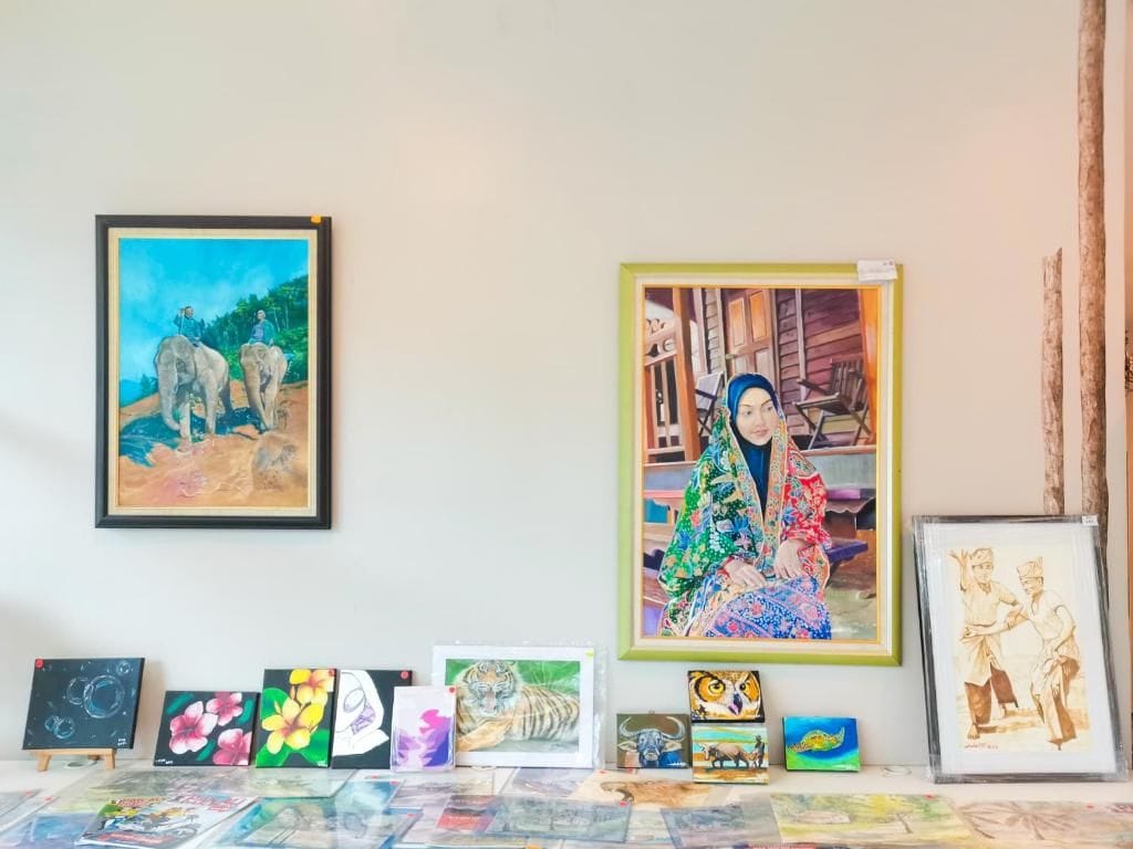Eine Wand mit verschiedenen gerahmten Kunstwerken, darunter ein Elefantenbild und eine farbenfrohe Figur.