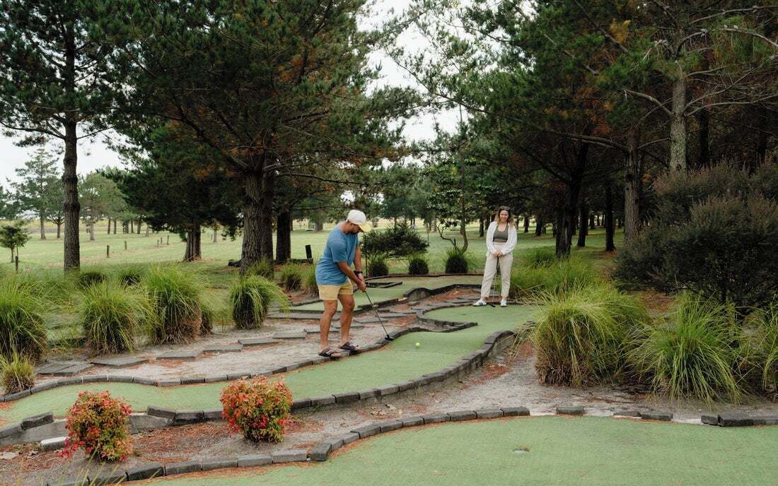 Um campo de mini golfe com superfície verde para tacadas, cercado por árvores e arbustos.
