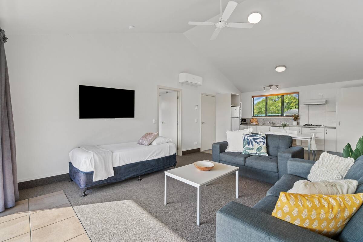 A modern living room with a bed, sofa, and coffee table. There is a kitchen in the background with a window.