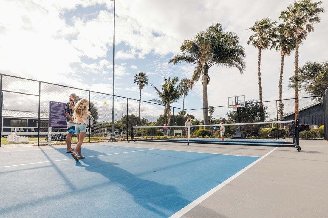 En udendørs tennisbane med net, omgivet af hegn og palmer.