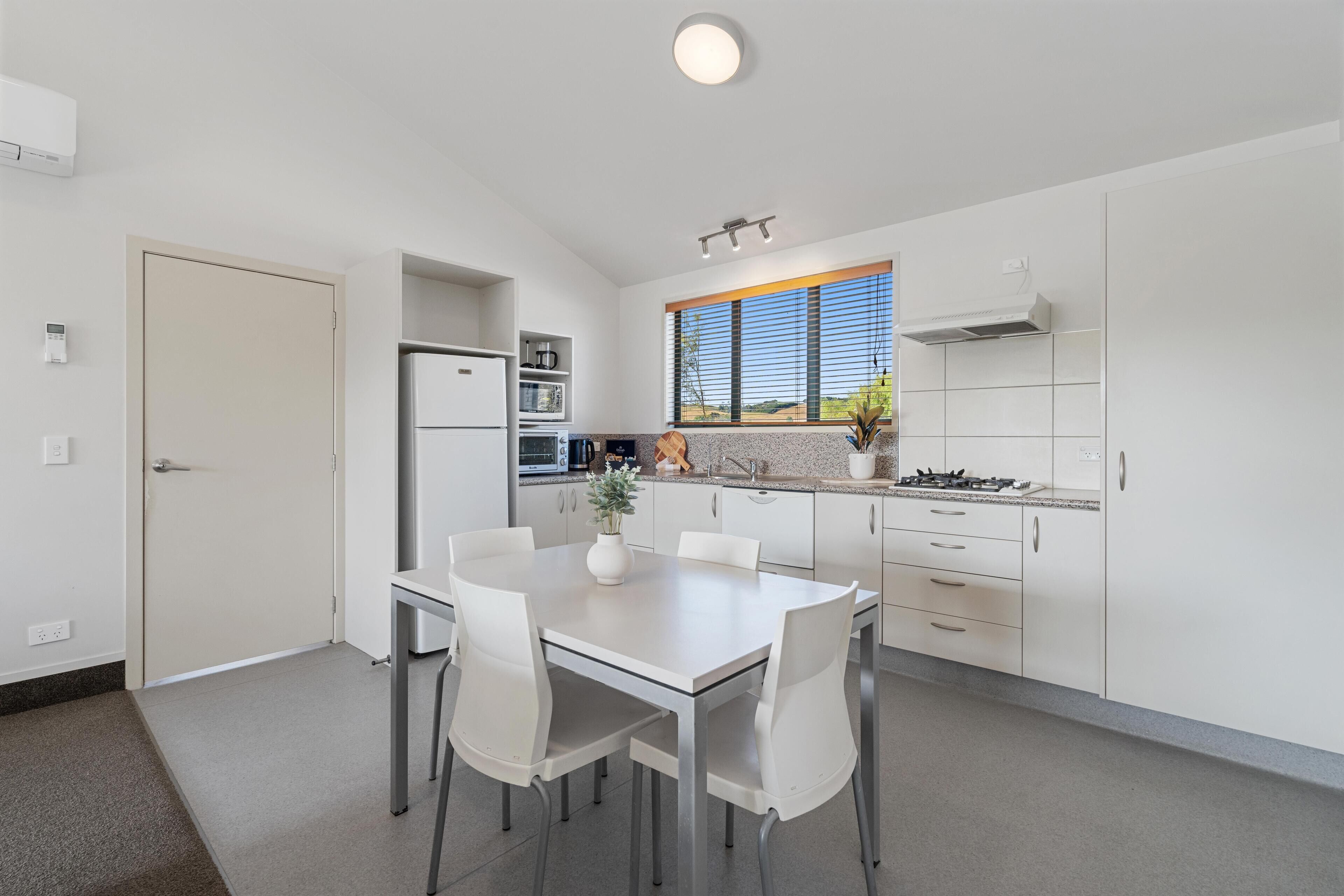 A modern kitchen with white cabinetry, a dining table with chairs, a built-in oven, and a window with a view of the outdoors.