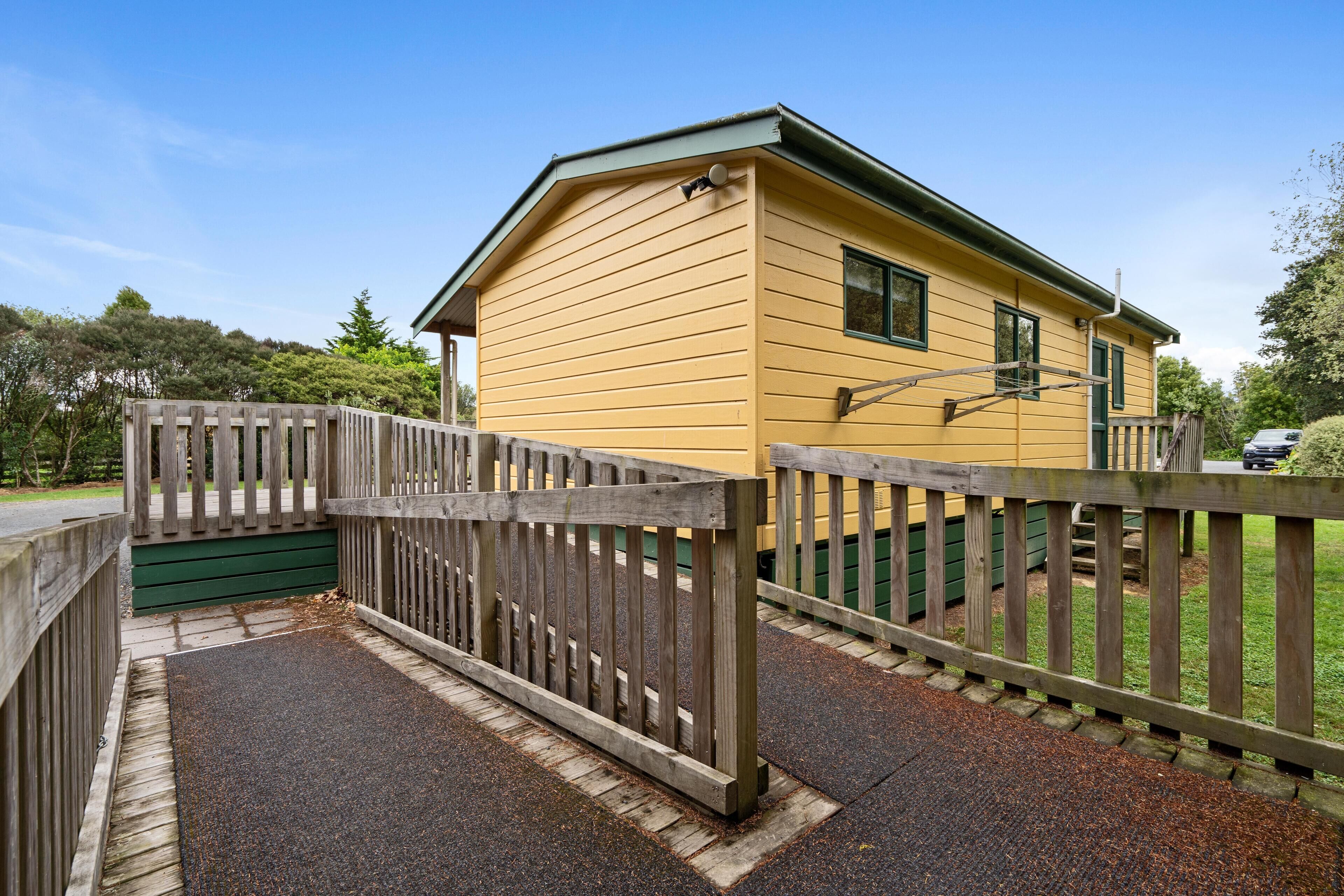 A yellow building with a green roof, a wooden fence, and a gravel driveway.