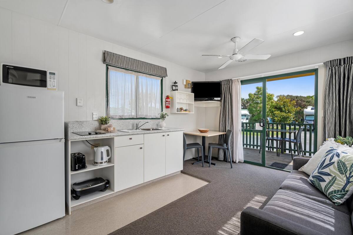 A compact kitchen with a microwave, toaster, and kettle, a dining area with a table and chairs, and a sofa with a view of the outdoors.