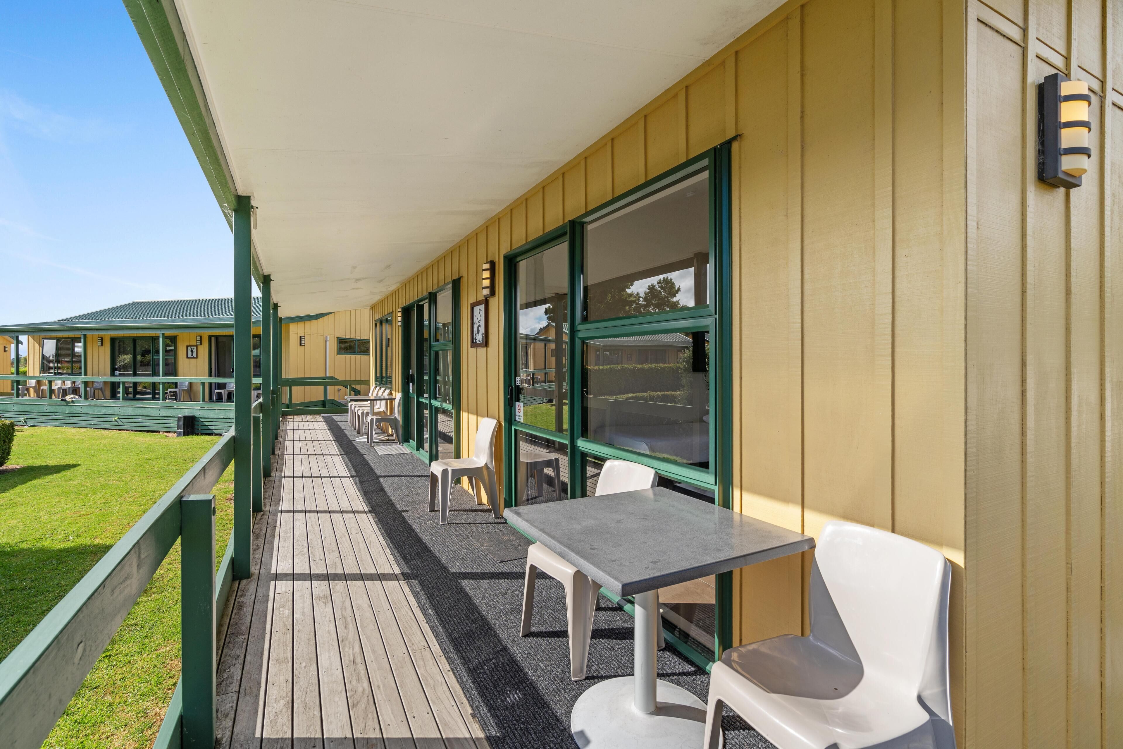 Un espacio de estar al aire libre cubierto con mesas y sillas, una terraza de madera y un edificio con grandes ventanales y techo verde.