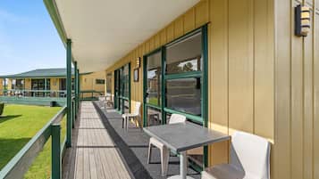 Un espacio de estar al aire libre cubierto con mesas y sillas, una terraza de madera y un edificio con grandes ventanales y techo verde.
