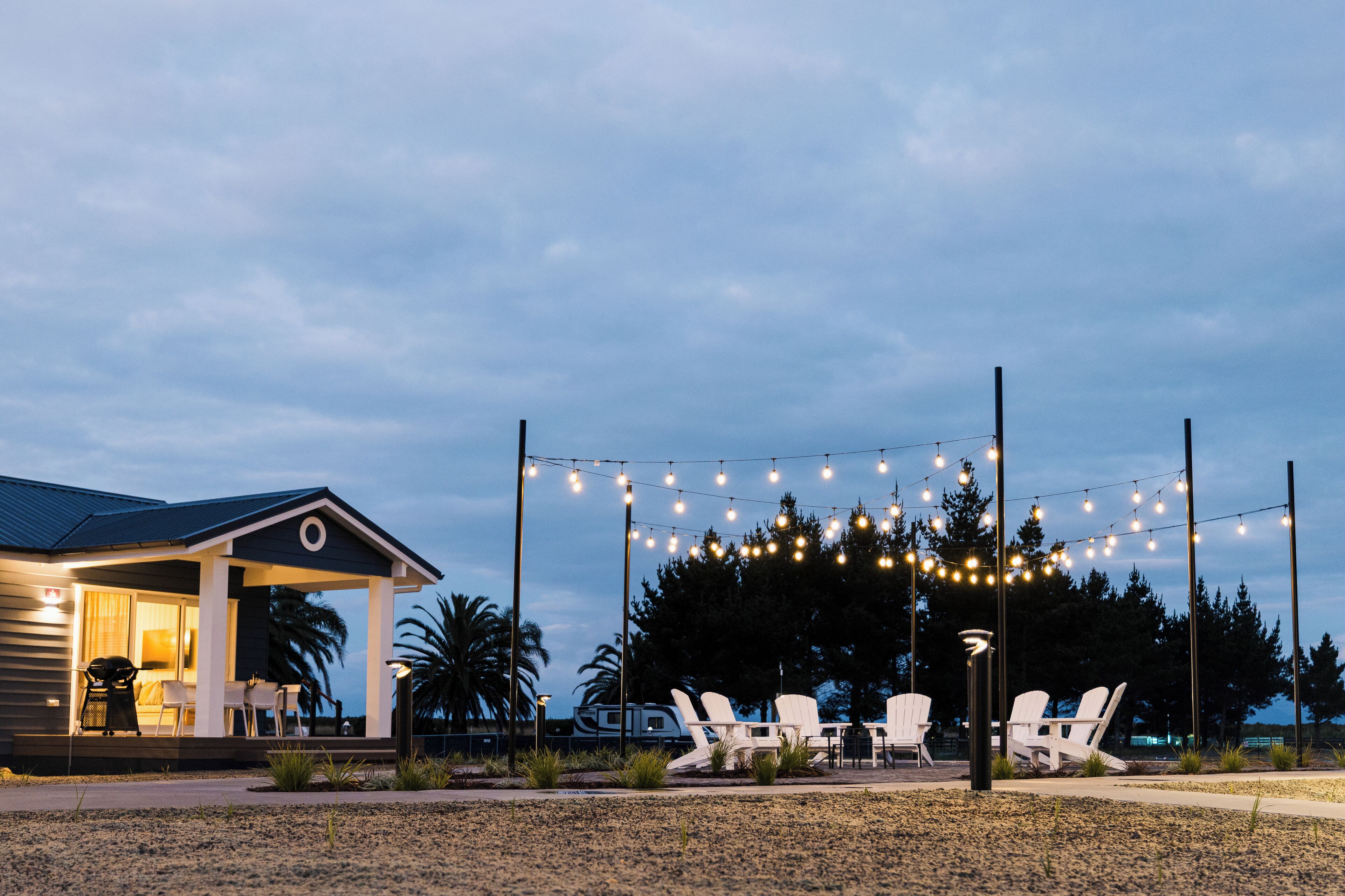 A covered outdoor seating area with string lights, white chairs, and a small building with a porch.