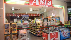 Uma loja de conveniência com um grande letreiro 'Asia', prateleiras repletas de lanches e uma máquina de vendas automática visível.