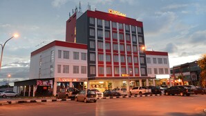 Um hotel de vários andares com fachada em vermelho e branco bem visível, vários espaços de estacionamento e um restaurante chamado "TRAVELER Lounge" no andar térreo.